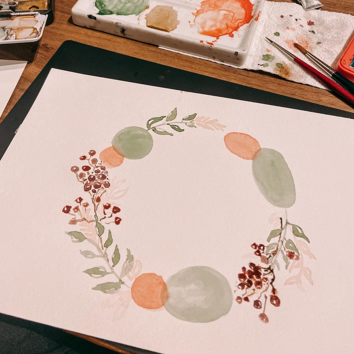 Blumenkranz in Aquarell gemalt auf Papier, Pinsel und Farben im Hintergrund – aquarell workshop, florale aquarellbilder.