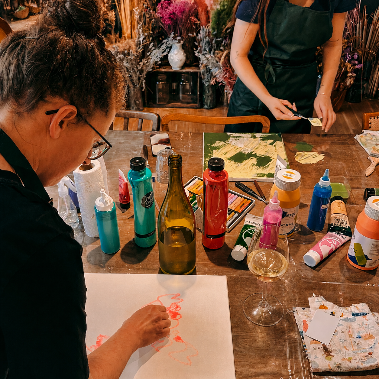 Zwei Personen malen mit Acrylfarben und Wachsmalstiften auf Leinwänden beim Abstract Painting Workshop von GRIMMS GARTEN Berlin. Dazu trinken sie ein Glas Wein.