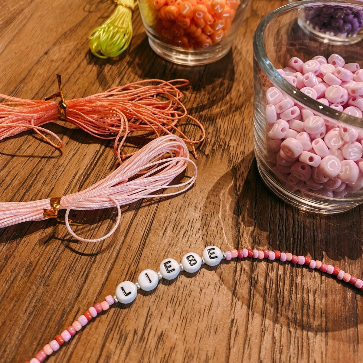 Perlenarmband mit Aufschrift „Liebe“ auf pinken Schnüren, Perlen im Hintergrund, kreativer DIY Schmuckworkshop bei Grimms Garten Berlin