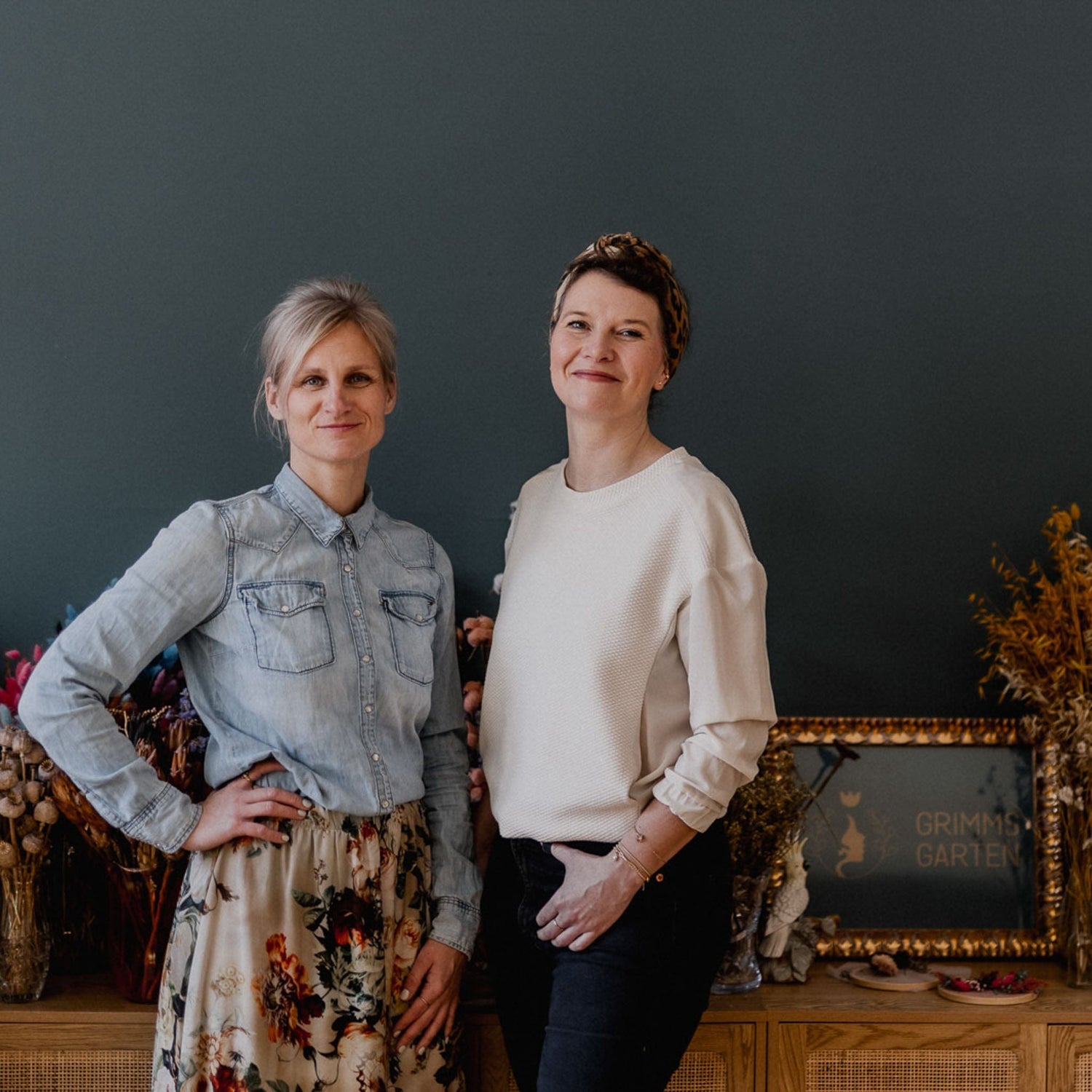 Zwei Frauen stehen vor dunkler Wand mit Grimms Garten Logo im Hintergrund, kreative Workshop-Atmosphäre in Berlin