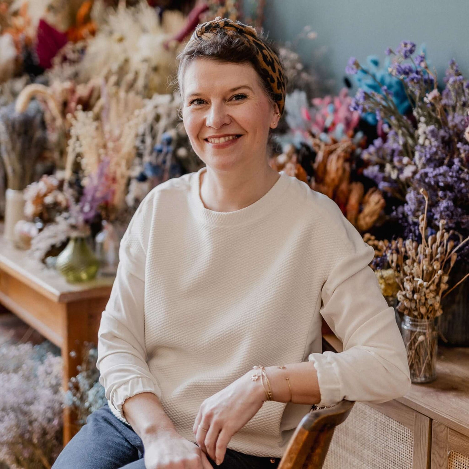 Frau mit Haarband sitzt lächelnd auf Holzstuhl vor Trockenblumen beim kreativen Workshop in Grimms Garten Berlin