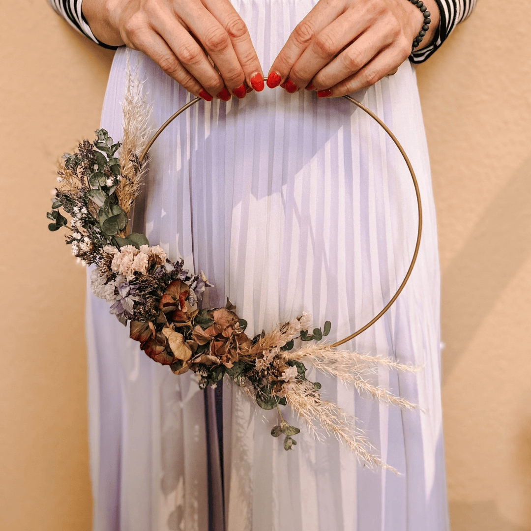 Hände halten bunten Trockenblumenring vor gelber Wand – kreative Schmuck Workshops im Grimms Garten