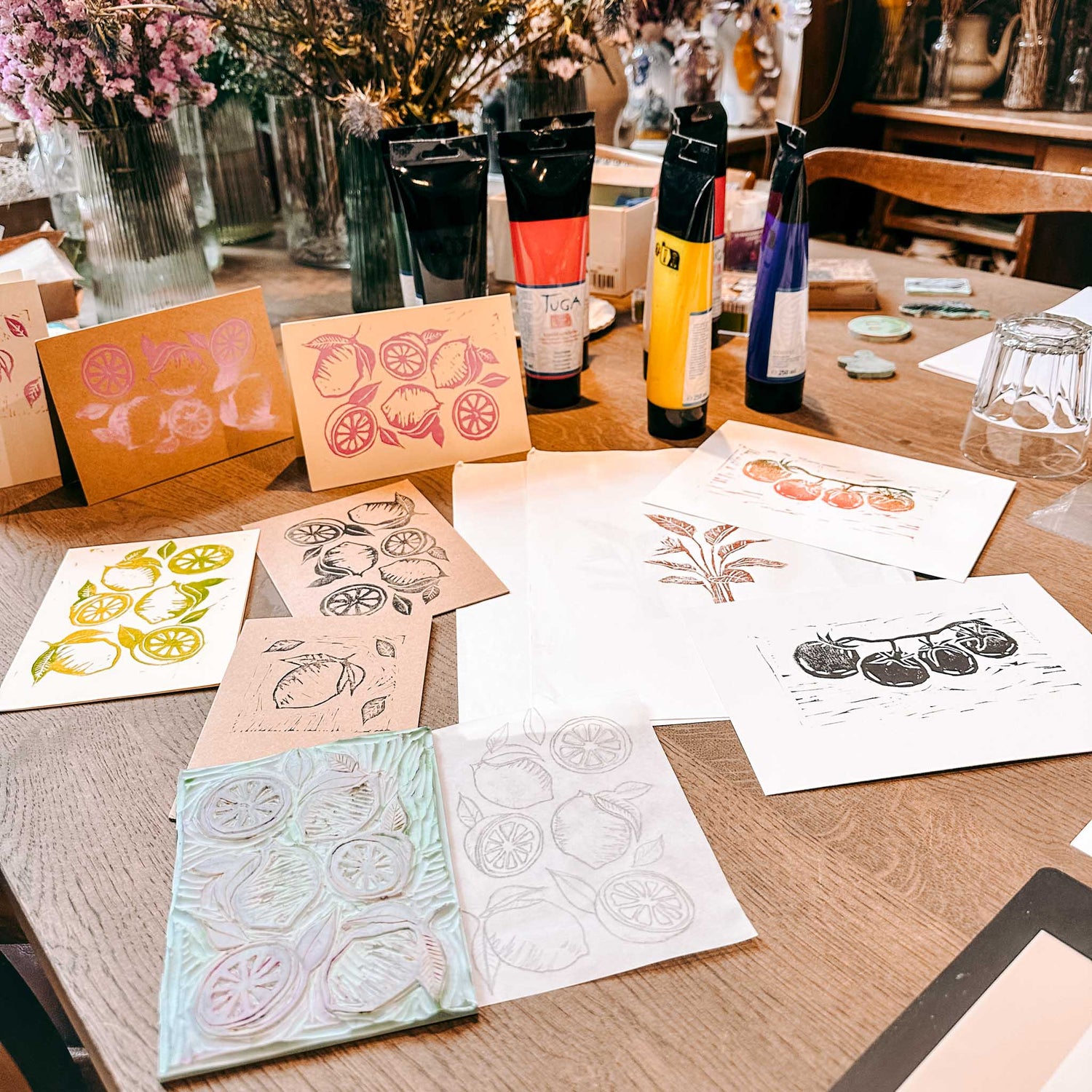 Mehrere fertige Drucke auf Tisch verteilt mit Farbtuben im Hintergrund – Linolschnitt, Linoldruck, DIY Workshop.