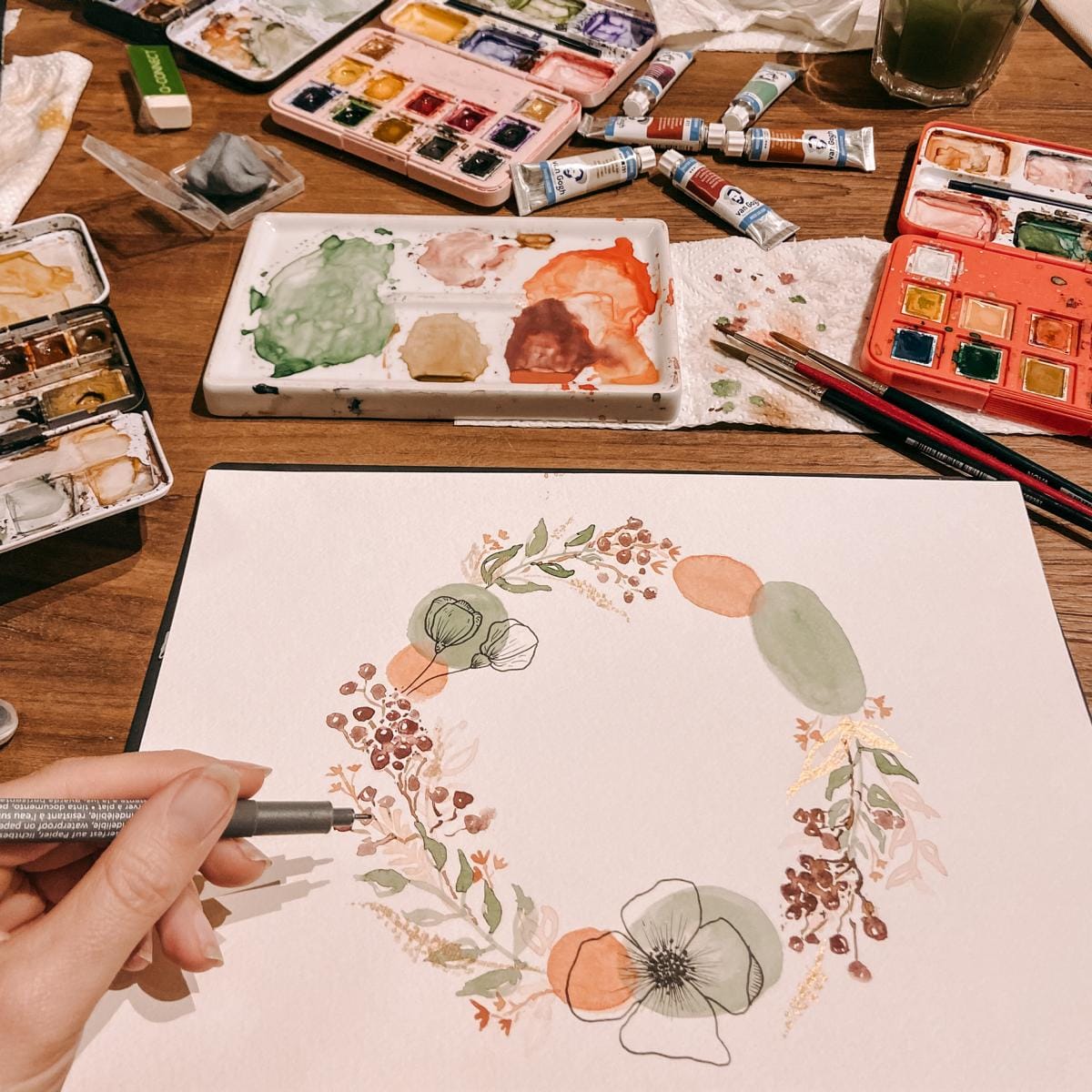 Blumenkranz auf Tisch mit Farben und Pinseln, Hand hält Fineliner – aquarell workshop, florale aquarellbilder, mixed media aquarell.