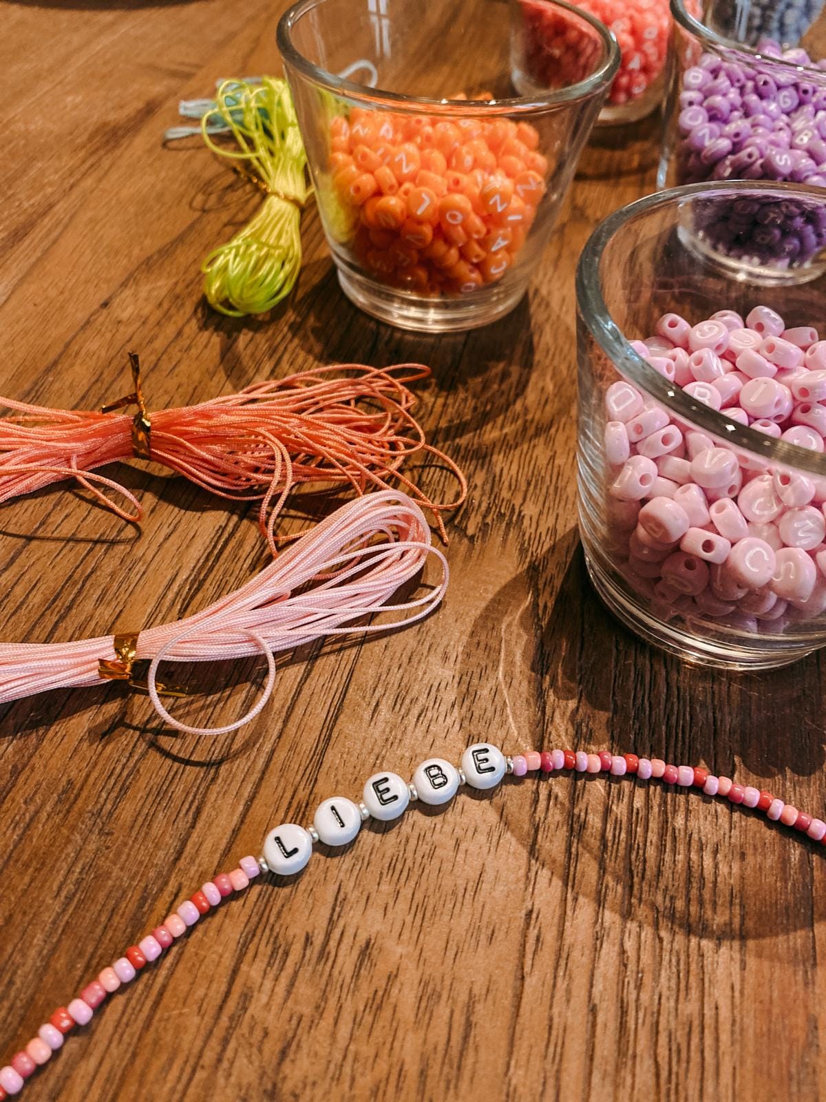 Perlenarmband mit Schriftzug „Liebe“ in Rosa und Rot auf dem Tisch, dahinter bunte Perlen und Bänder beim DIY Perlenschmuck Workshop von GRIMMS GARTEN in Berlin