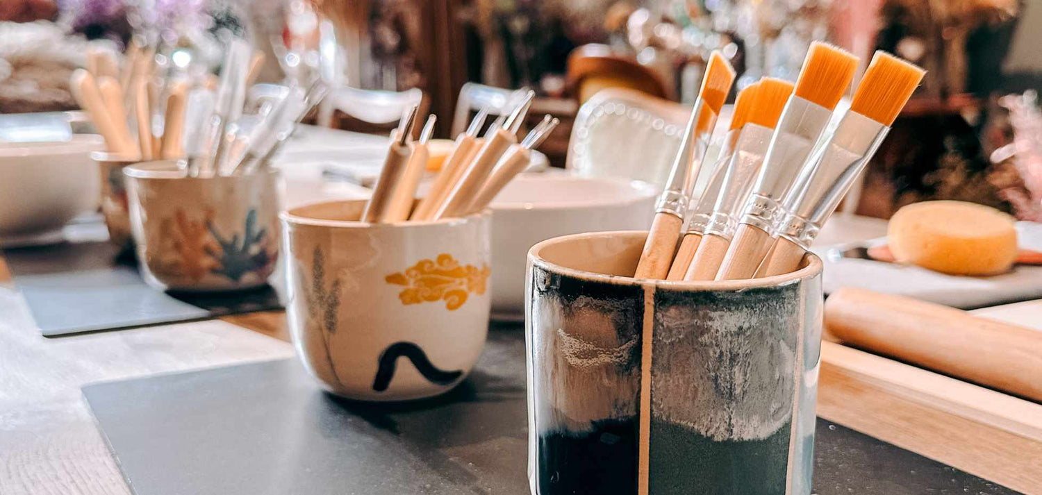 Handbemalte Gefäße mit Pinseln darin, auf einem Tisch hintereinander aufgereiht.