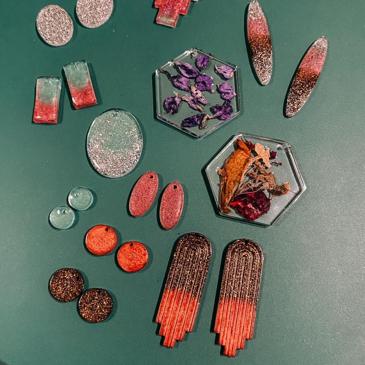 Verschiedene Resin Schmuckteile auf blauem Hintergrund – DIY Epoxidharz Schmuck mit Glitzer und Blumen.