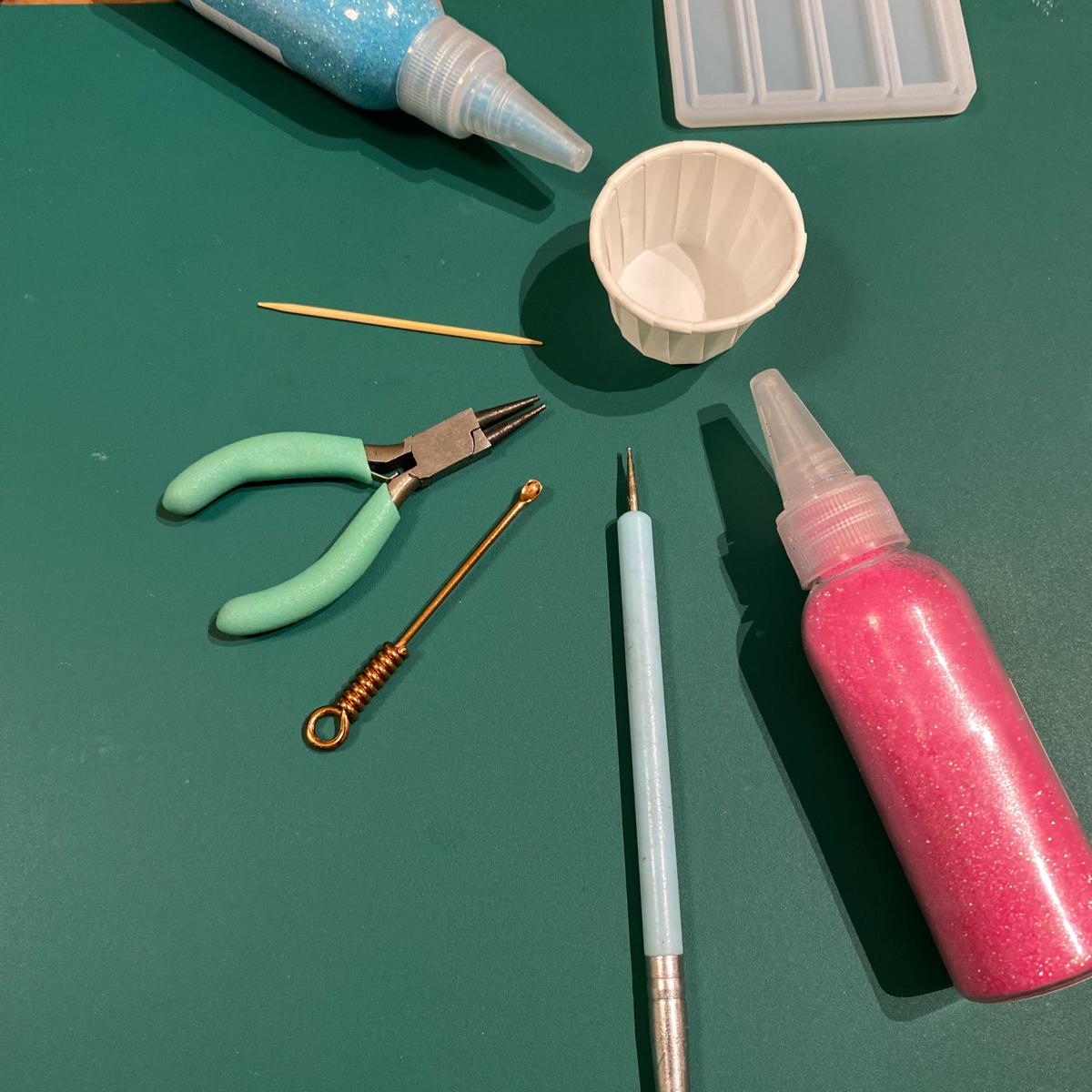 Glitzer, Zange und Materialien auf blauem Hintergrund – Resin Schmuck Workshop für kreative Designs.