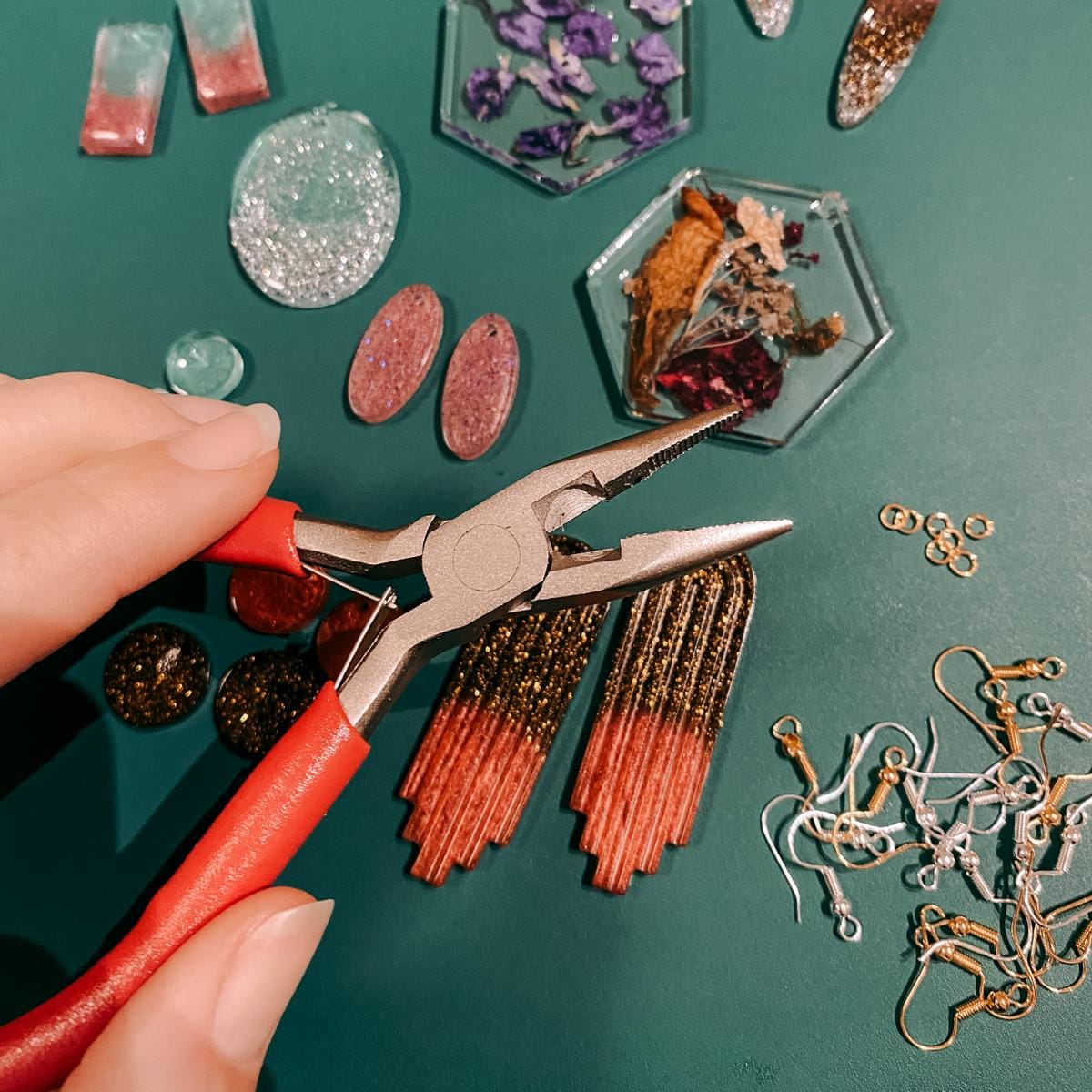 Hand hält Zange vor Resin Schmuckteilen – kreativer Epoxidharz Schmuckworkshop mit Ohrringen und Anhängern.