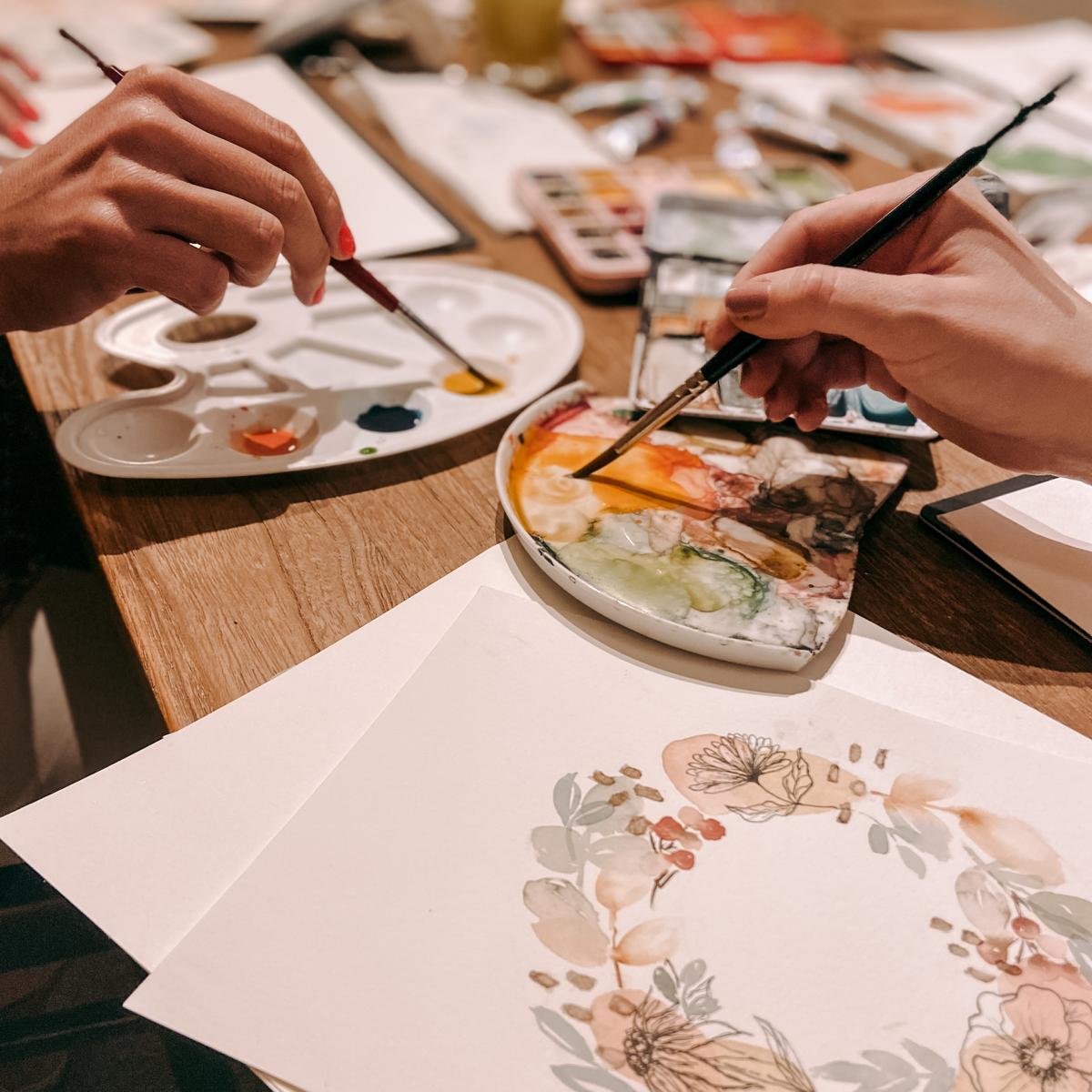 Zwei Hände tauchen Pinsel in Farbe, Bild liegt auf Tisch – Aquarell Workshop, Watercolour, DIY Karten, auch für Firmenevents