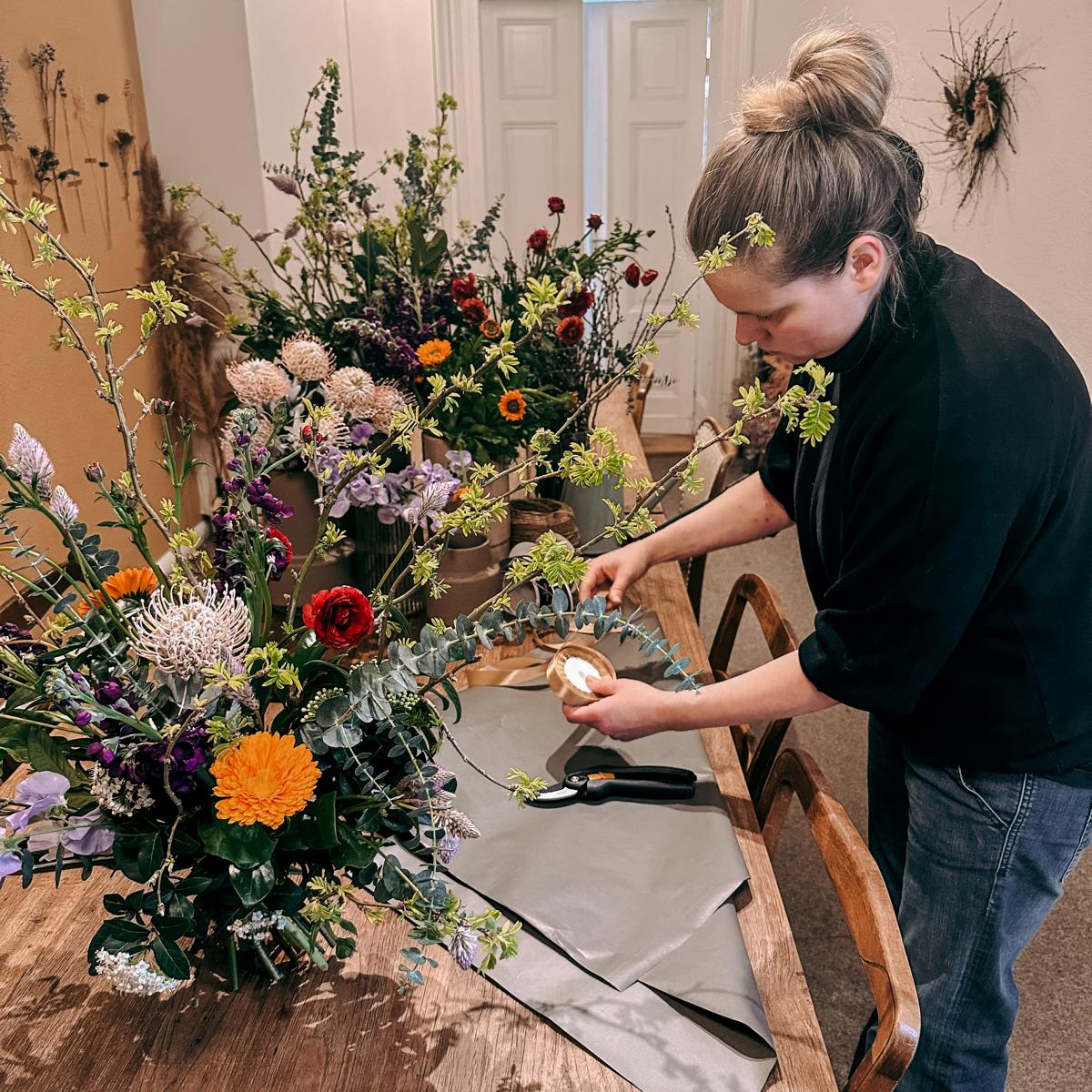 Frau erstellt Blumenstrauß an Tisch mit mehreren Sträußen – kreativer Workshop, Teamevent geeignet.