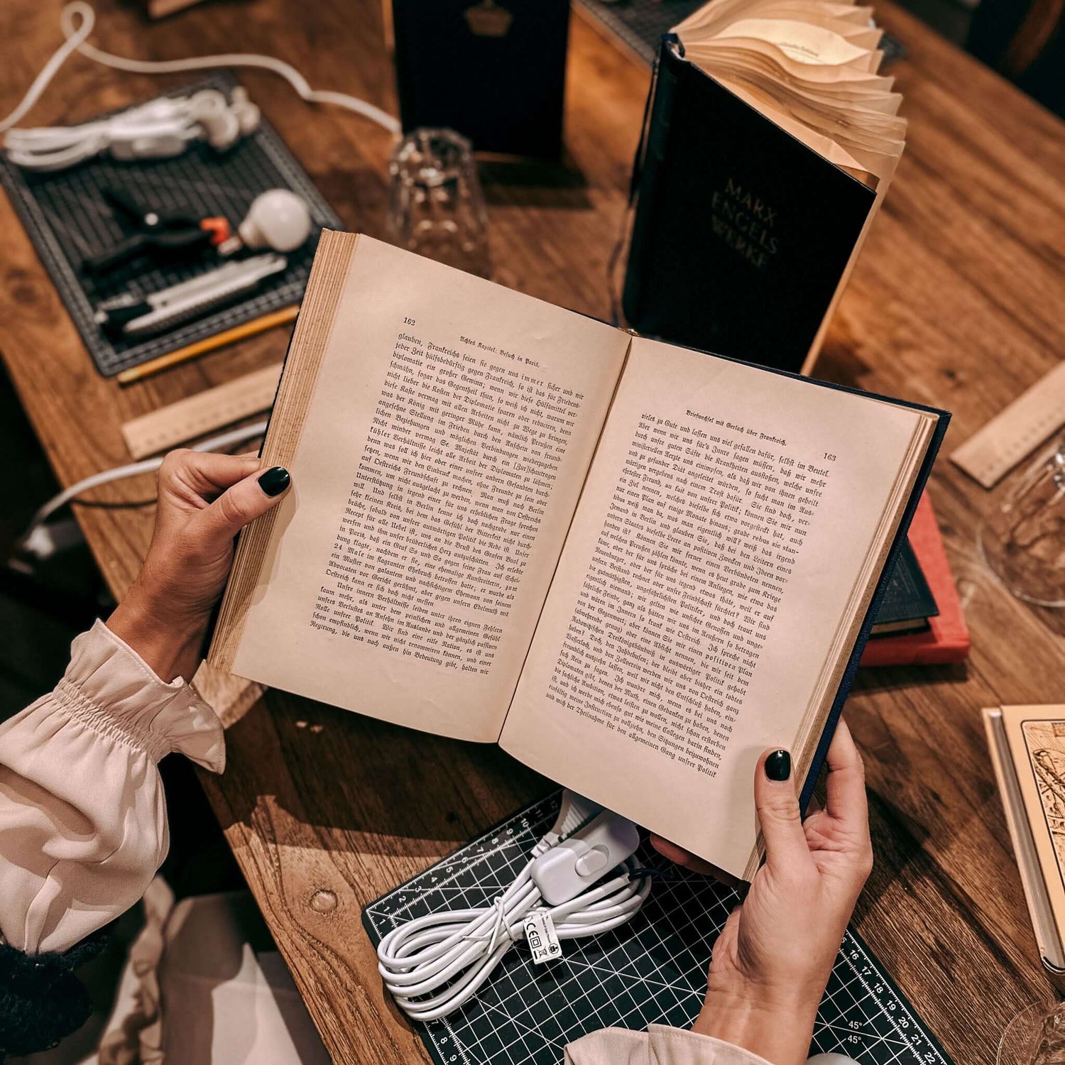 Zwei Hände halten Buch offen über Tisch mit Materialien – upcycling buchlampe, diy workshop, kreative lampe. Auch für Firmenevents.