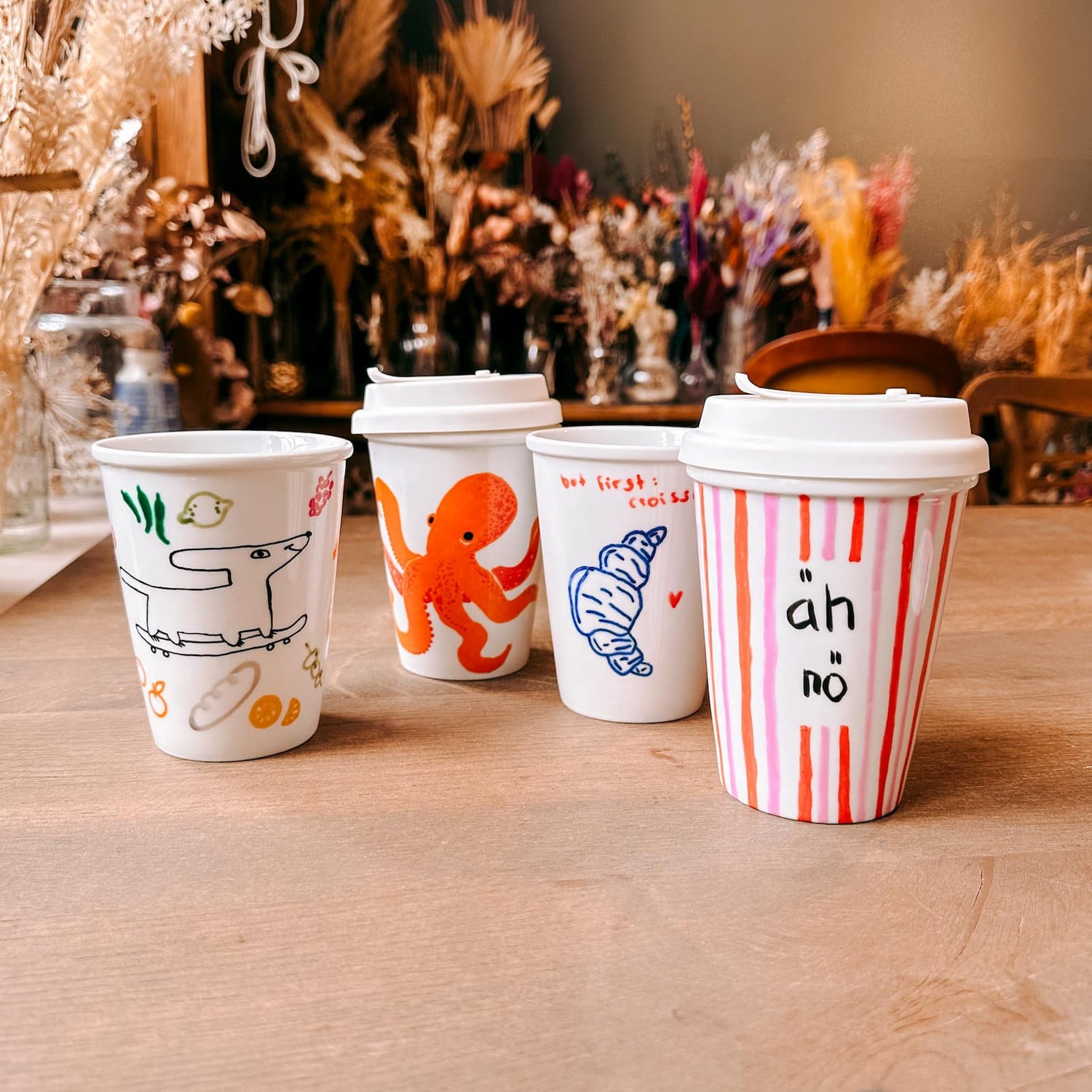 Vier Keramik To-Go Becher mit unterschiedlichen Motiven und Farben auf dem Tisch beim Keramik bemalen Workshop von GRIMMS GARTEN, Kreativkurs in Berlin