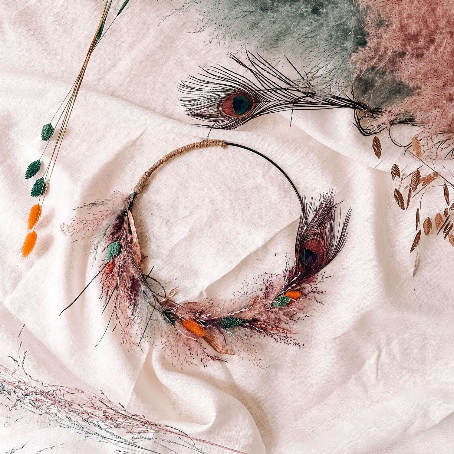 Bunter Trockenblumenring Pfauenauge auf weißem Tuch mit Federn und Trockenblumen im Hintergrund.
