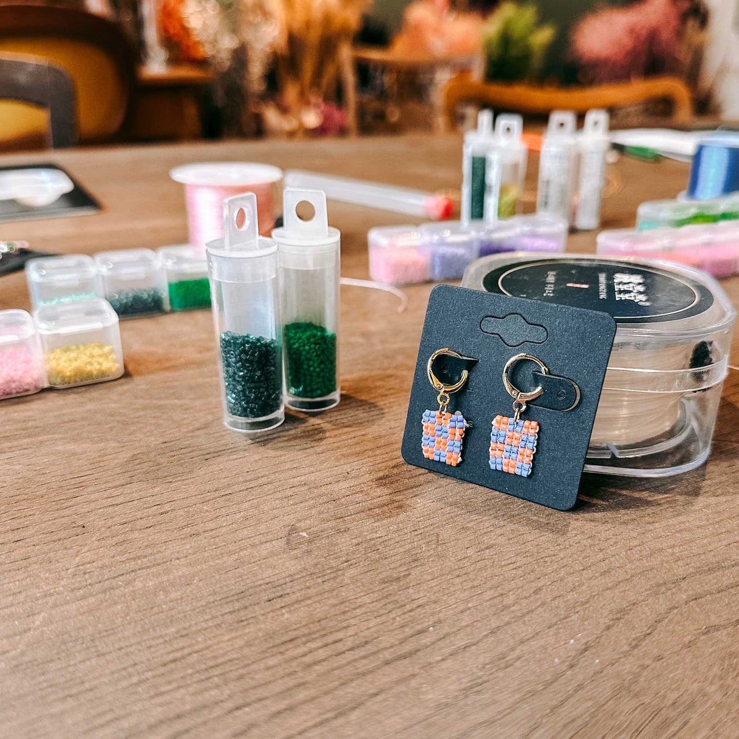 Fertiges Paar Ohrringe in Blau und Orange mit bunten Perlen im Hintergrund beim DIY Perlenschmuck Workshop von GRIMMS GARTEN, Kreativkurs in Berlin