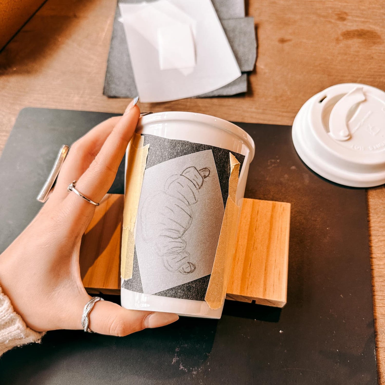 Hand hält Keramik To-Go Becher mit aufgeklebter Croissant-Skizze als Vorlage beim Keramik bemalen Workshop von GRIMMS GARTEN, Kreativkurs in Berlin