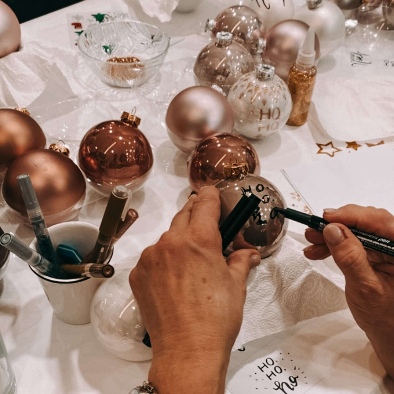 Zwei Hände bemalen Weihnachtskugel mit Stift – weitere Kugeln im Hintergrund.