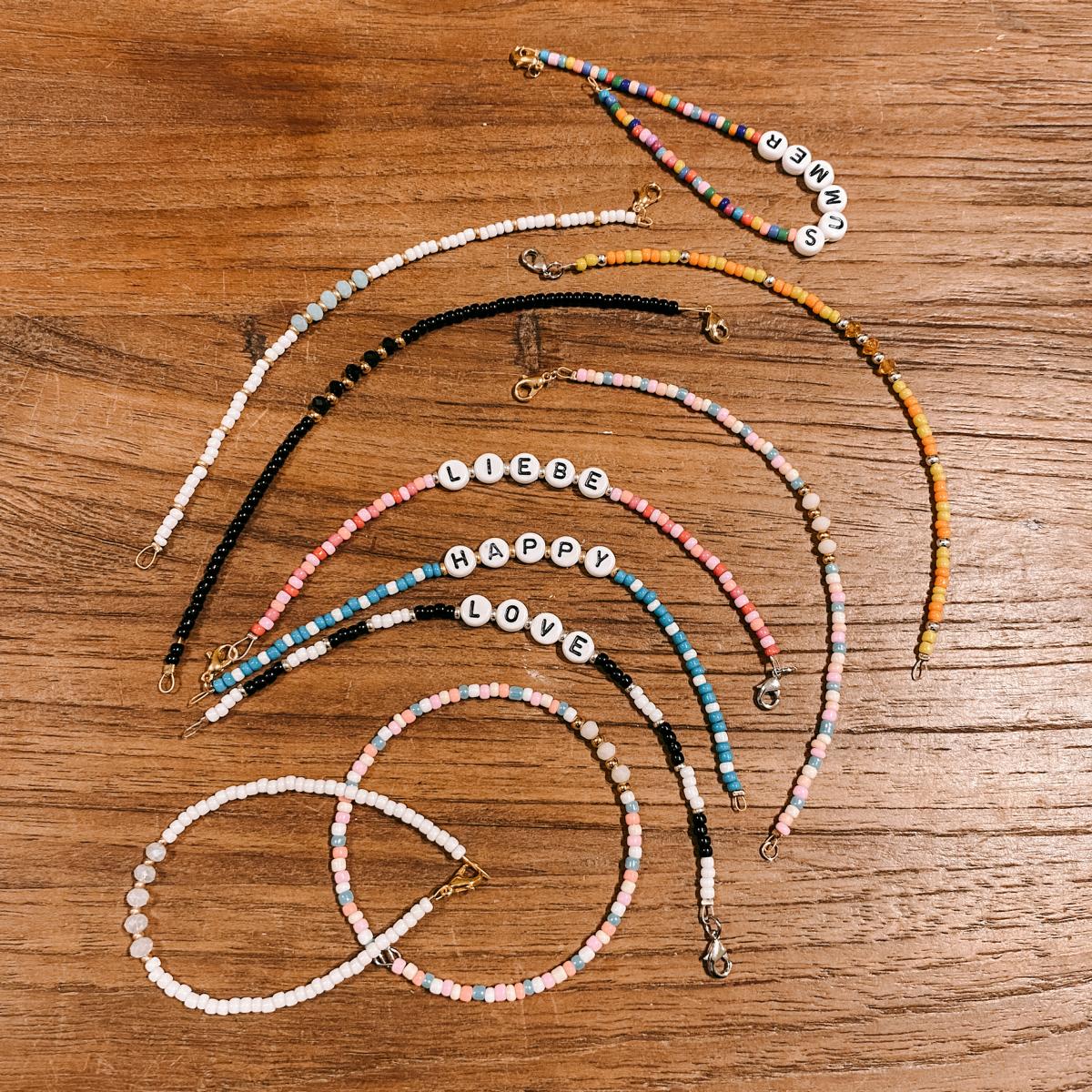 Verschiedene Armbänder mit Schriftzügen „Liebe, Happy, Love“ auf Holztisch – DIY Perlenschmuck Workshop.