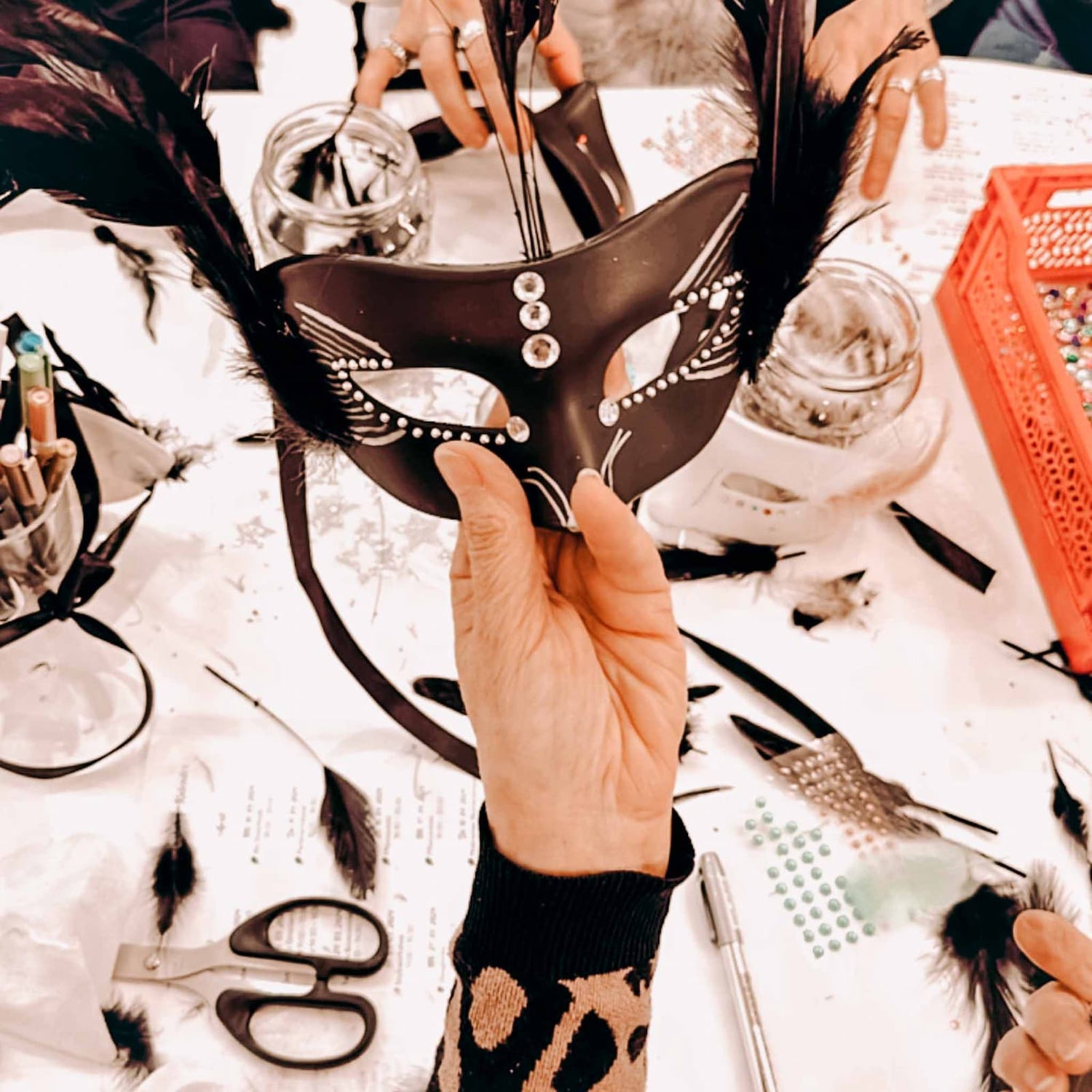 Hand hält schwarze Maske mit Federn und Glitzerelementen beim kreativen Masken Workshop von GRIMMS GARTEN, DIY Teamevent in Berlin