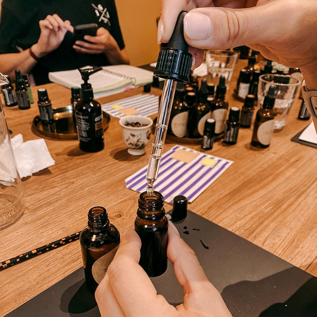 Hand hält Pipette über Fläschchen mit ätherischen Ölen im Hintergrund – diy parfum, ätherische öle, naturduft workshop, auch für Firmenevents.