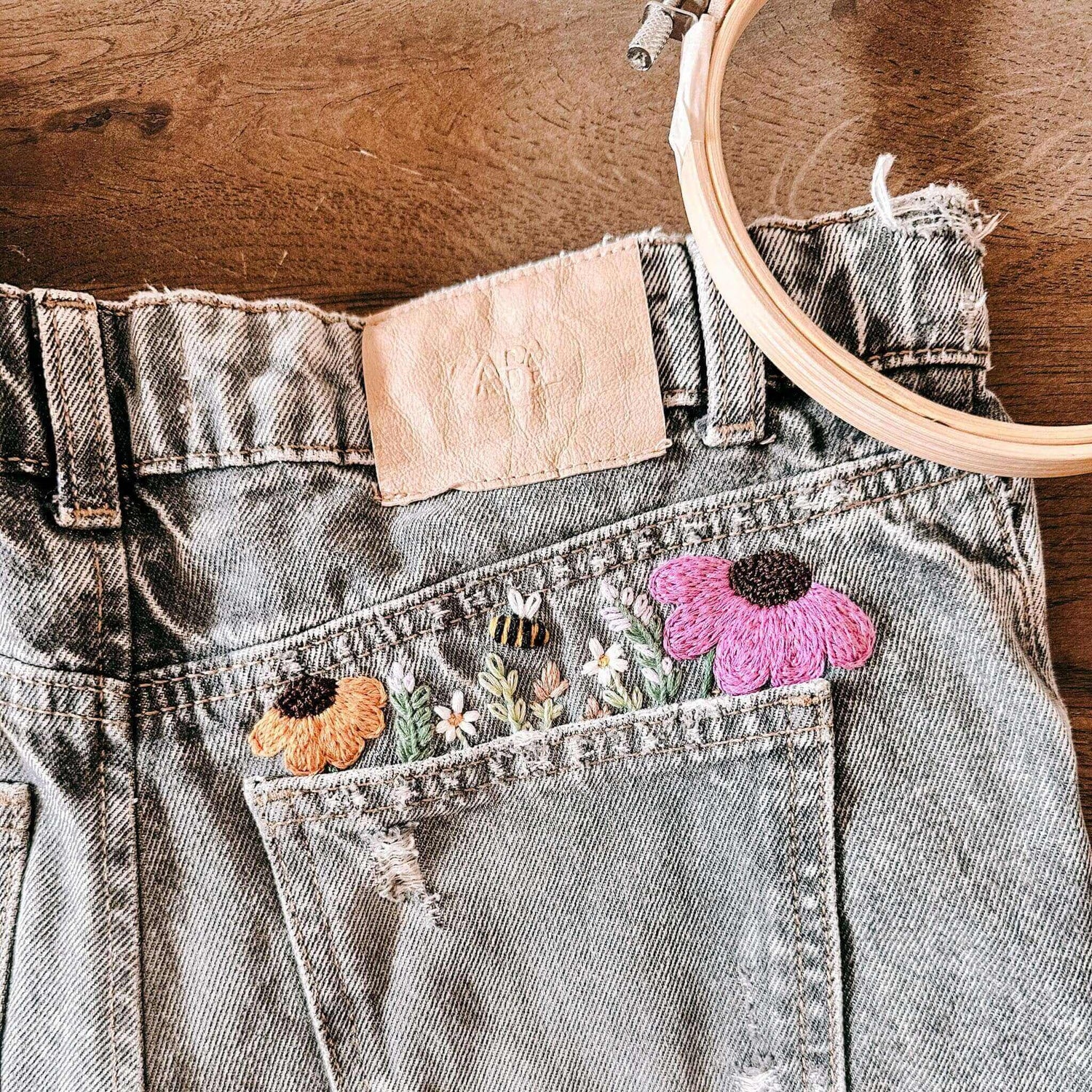 Graue Jeans mit bunten gestickten Blumen auf Holztisch – jeans besticken, handstickerei, nachhaltige kleidung, diy fashion.