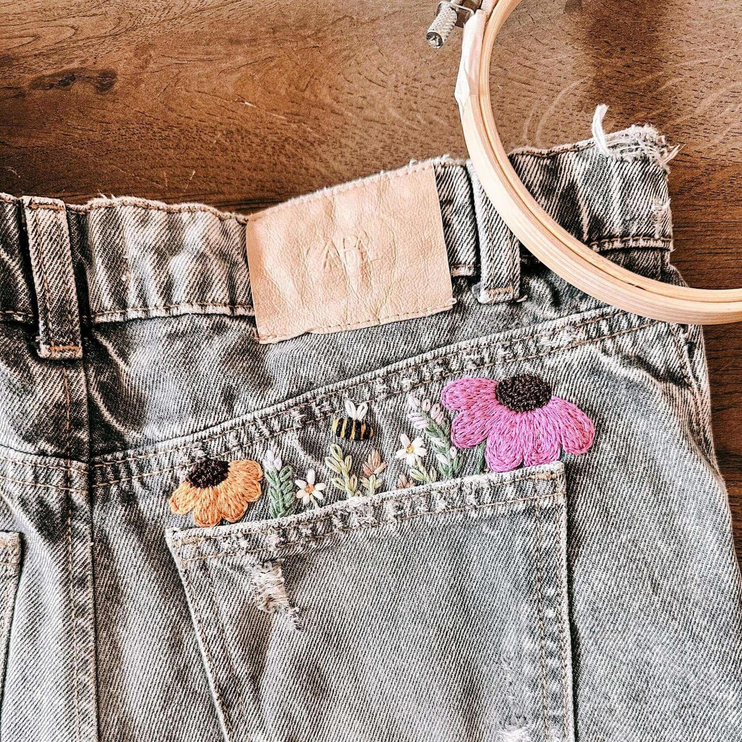 Graue Jeans mit bunten gestickten Blumen auf Holztisch – jeans besticken, handstickerei, nachhaltige kleidung, diy fashion.