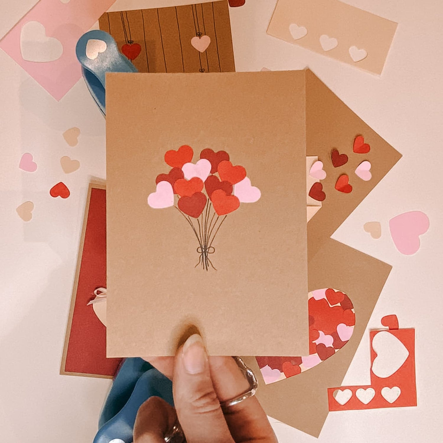 Hand hält Postkarte mit Blumenstrauß aus roten und rosa Herzen, im Hintergrund weitere gestaltete Karten beim kreativen Postkarten Workshop von GRIMMS GARTEN in Berlin