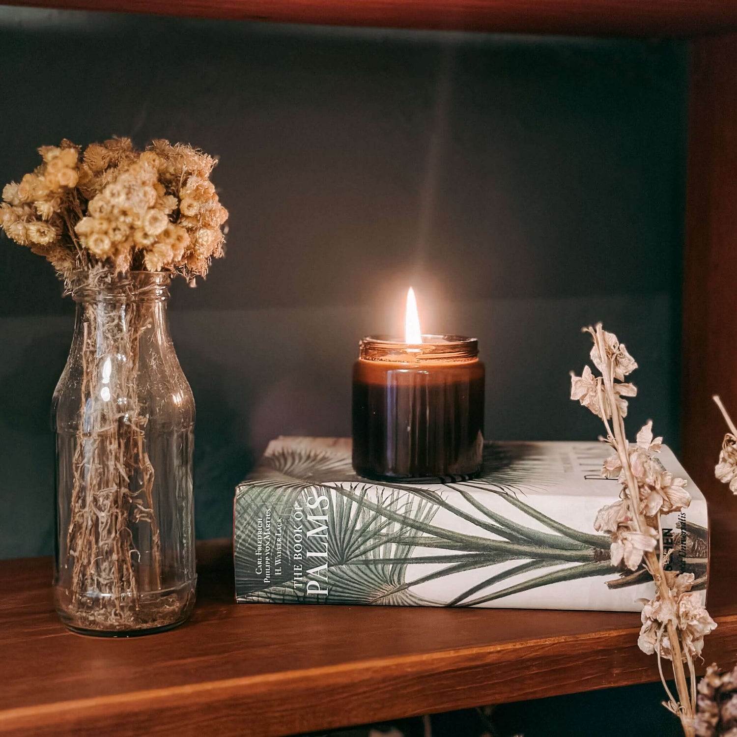 Brennende Duftkerze auf Buch mit Vase und Trockenblumen – stilvolle nachhaltige Deko.