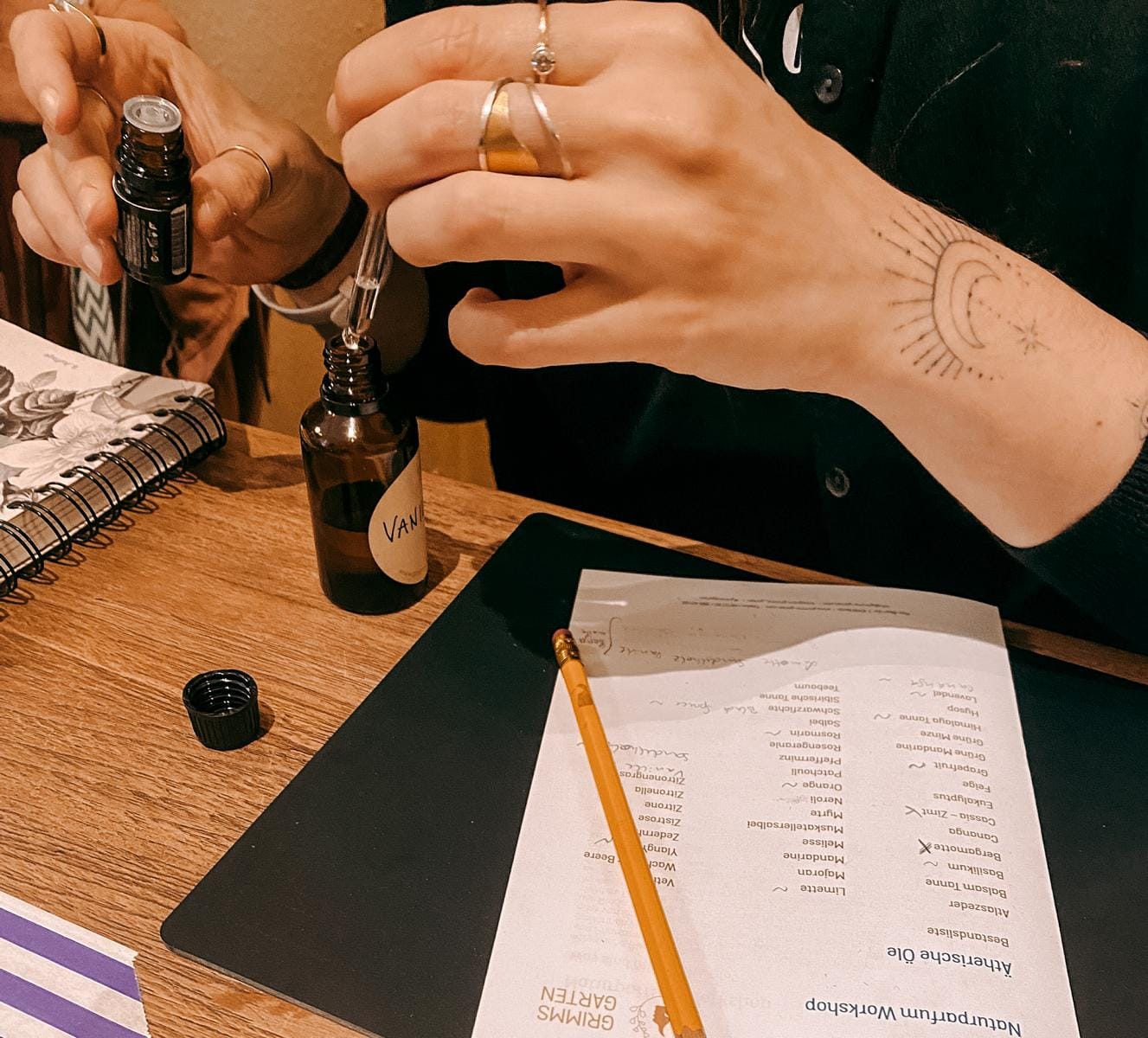 Hand tropft Flüssigkeit in kleine Flasche mit Zettel und Stift auf Tisch – naturparfum herstellen, aromatherapie workshop, diy duft, auch für Firmenevents.