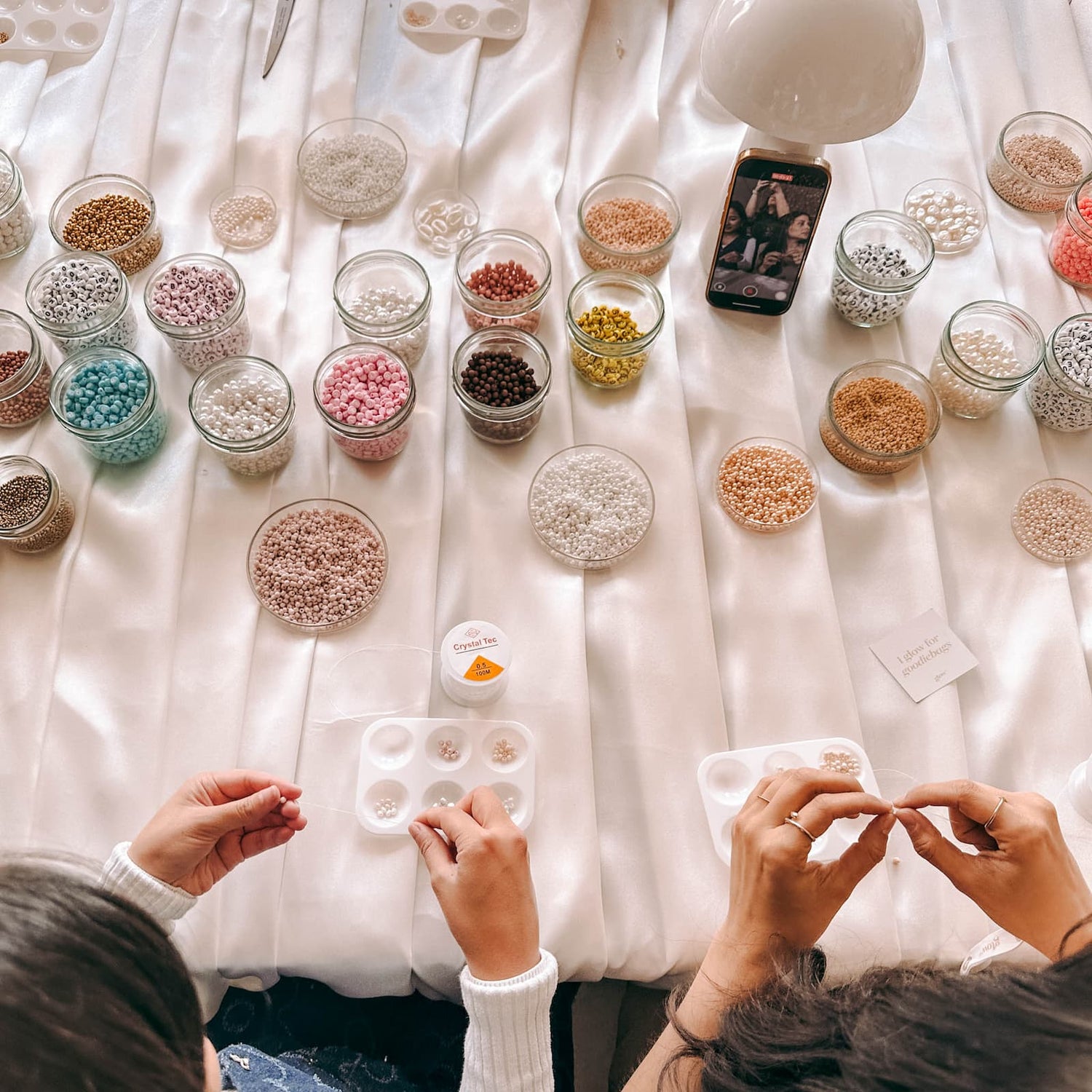 Großer Tisch mit Gläsern und Perlen, zwei Personen basteln Armbänder – DIY Perlenschmuck Workshop.