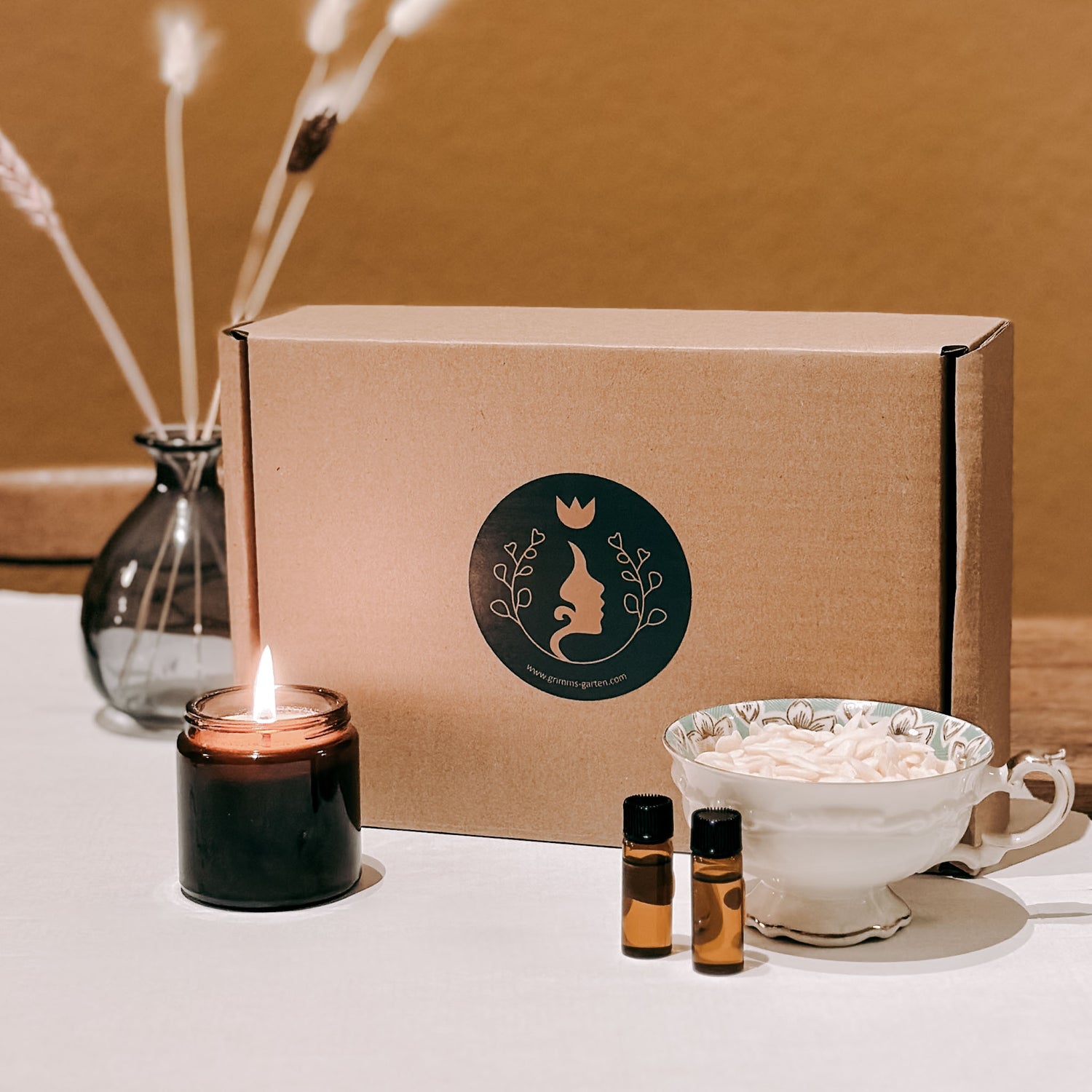 Brennende vegane Duftkerze neben Rapswachschips und DIY Box mit Grimms Garten Logo.