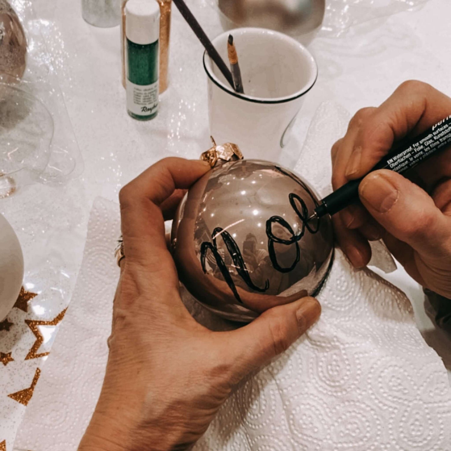 Zwei Hände schreiben „Noel“ auf Kugel – DIY Christbaumschmuck Workshop.