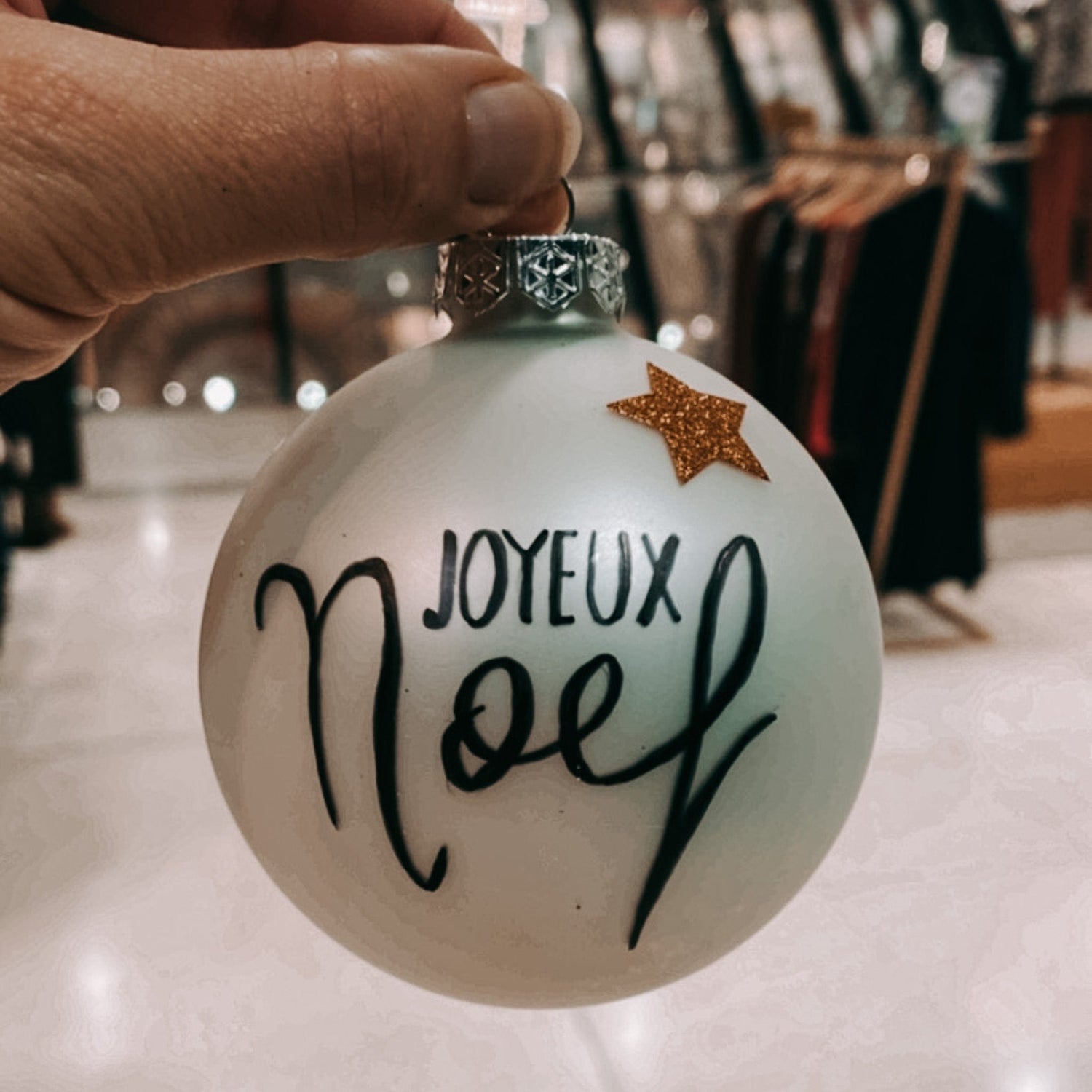 Hand hält weiße Weihnachtskugel hoch mit Aufschrift „Joyeux Noël“ – DIY Kugeln bemalen.