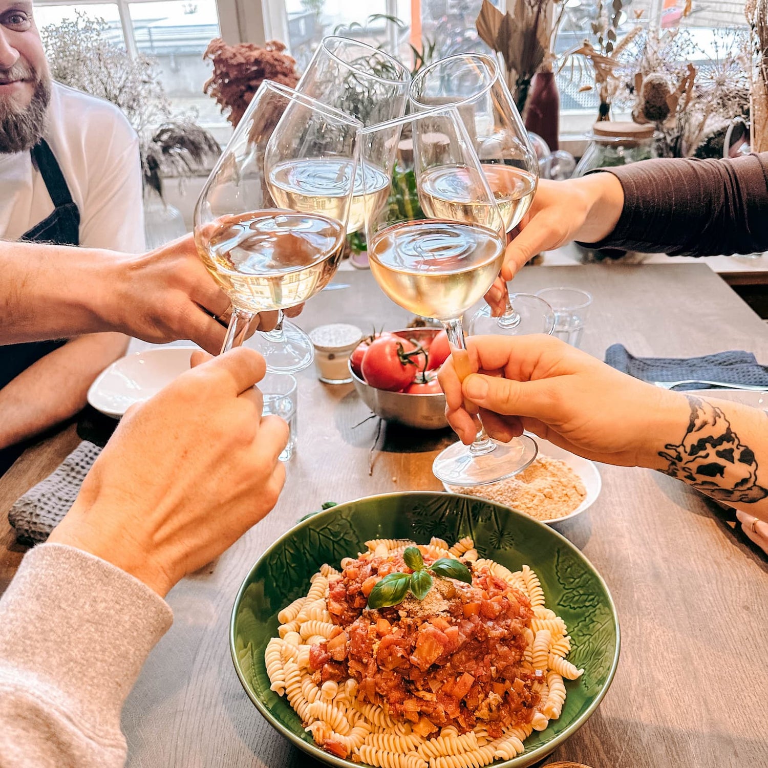 Vier Weingläser stoßen an, auf dem Tisch stehen Nudelgerichte beim Batch Cooking Workshop von GRIMMS GARTEN, Meal Prep Kochkurs in Berlin