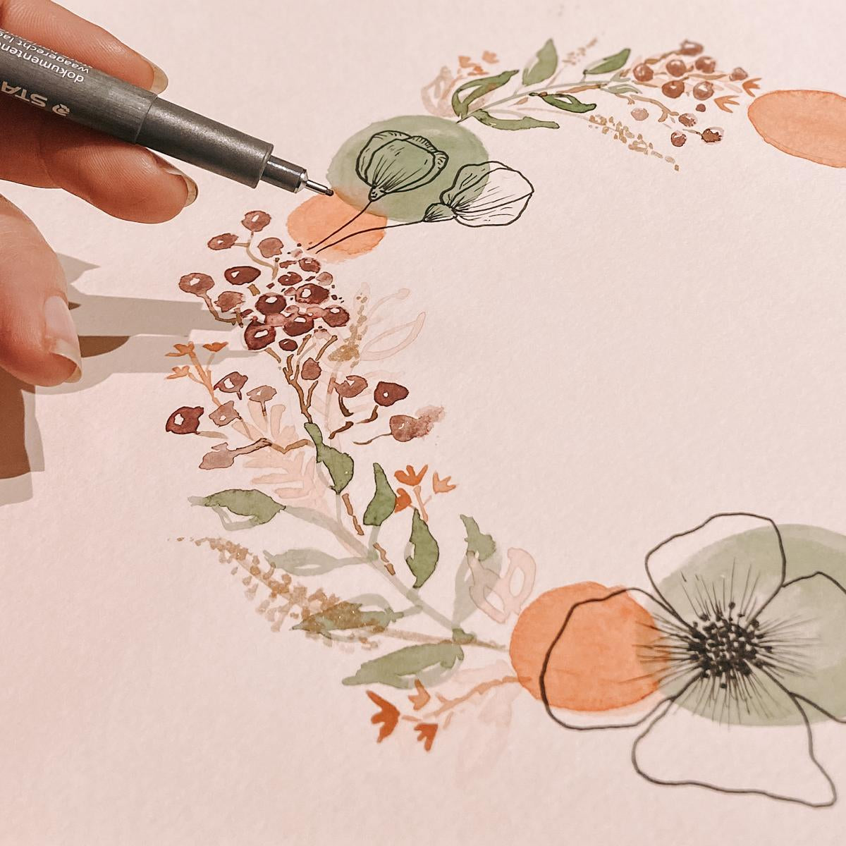 Hand zeichnet feine Linien mit Fineliner auf einen Aquarell-Blumenkranz – mixed media, aquarelltechnik, floral art.