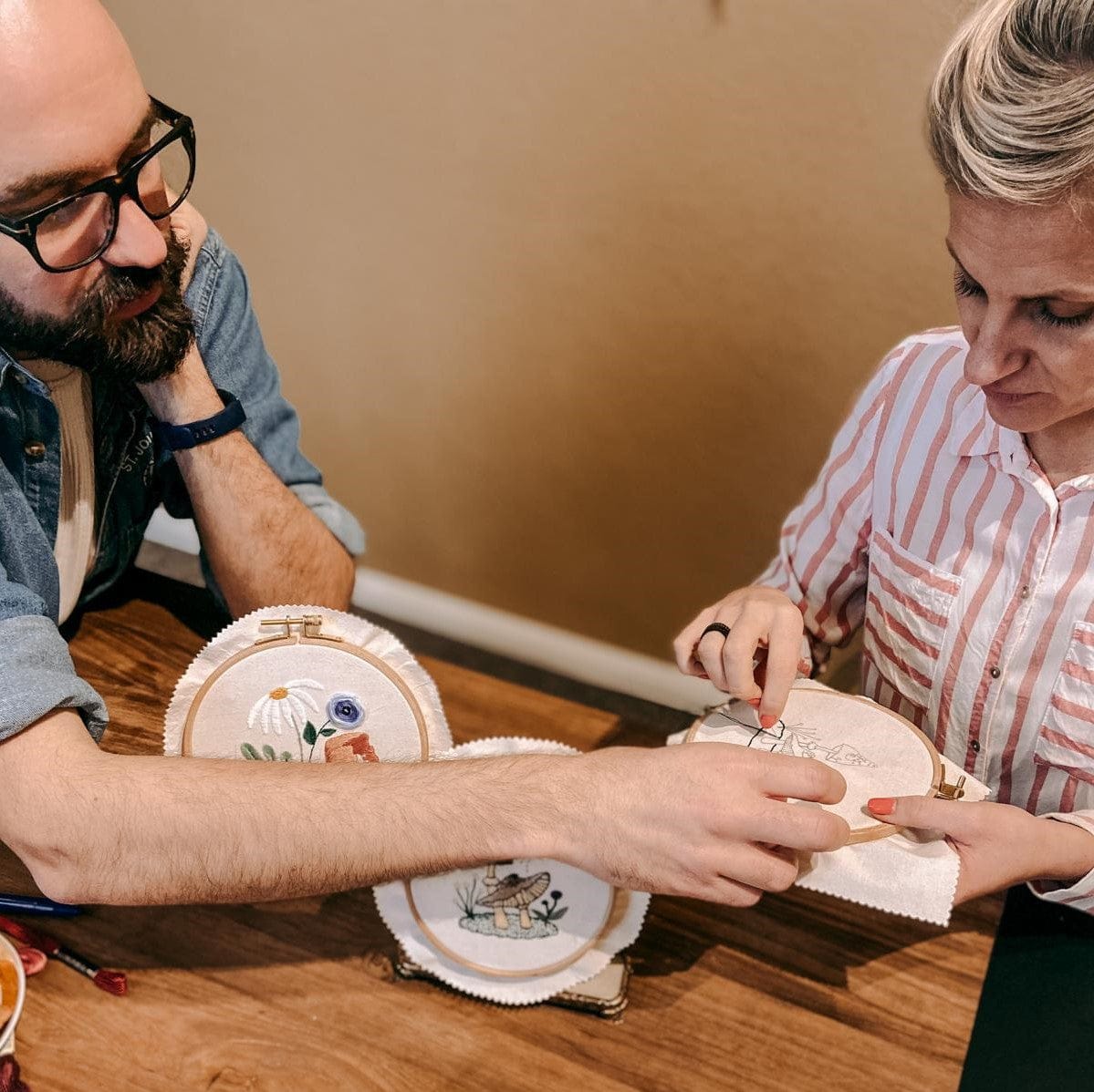 Zwei Personen bearbeiten Stickereien mit fertigem Blumenprojekt auf Tisch.