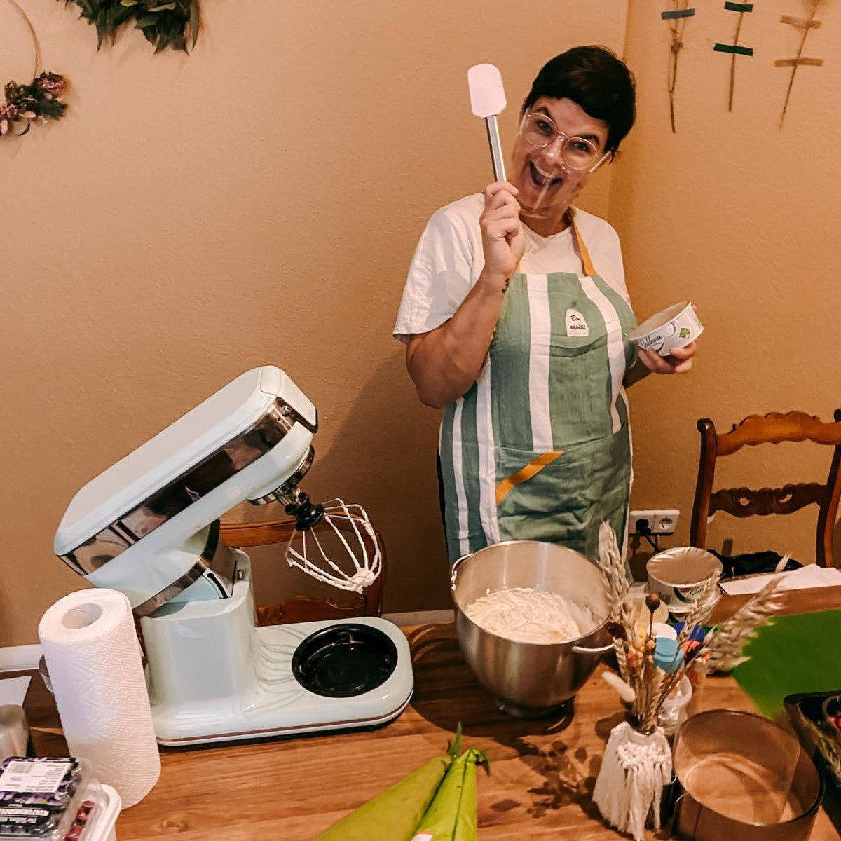 Frau hält Spatel vor Zutaten und KitchenAid – Backkurs Mini Drip Cake.