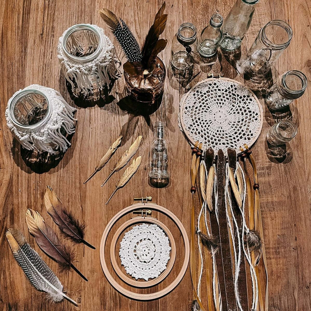 Verschiedene Boho-Deko wie Makramee, Traumfänger und Gläser auf Holztisch arrangiert.