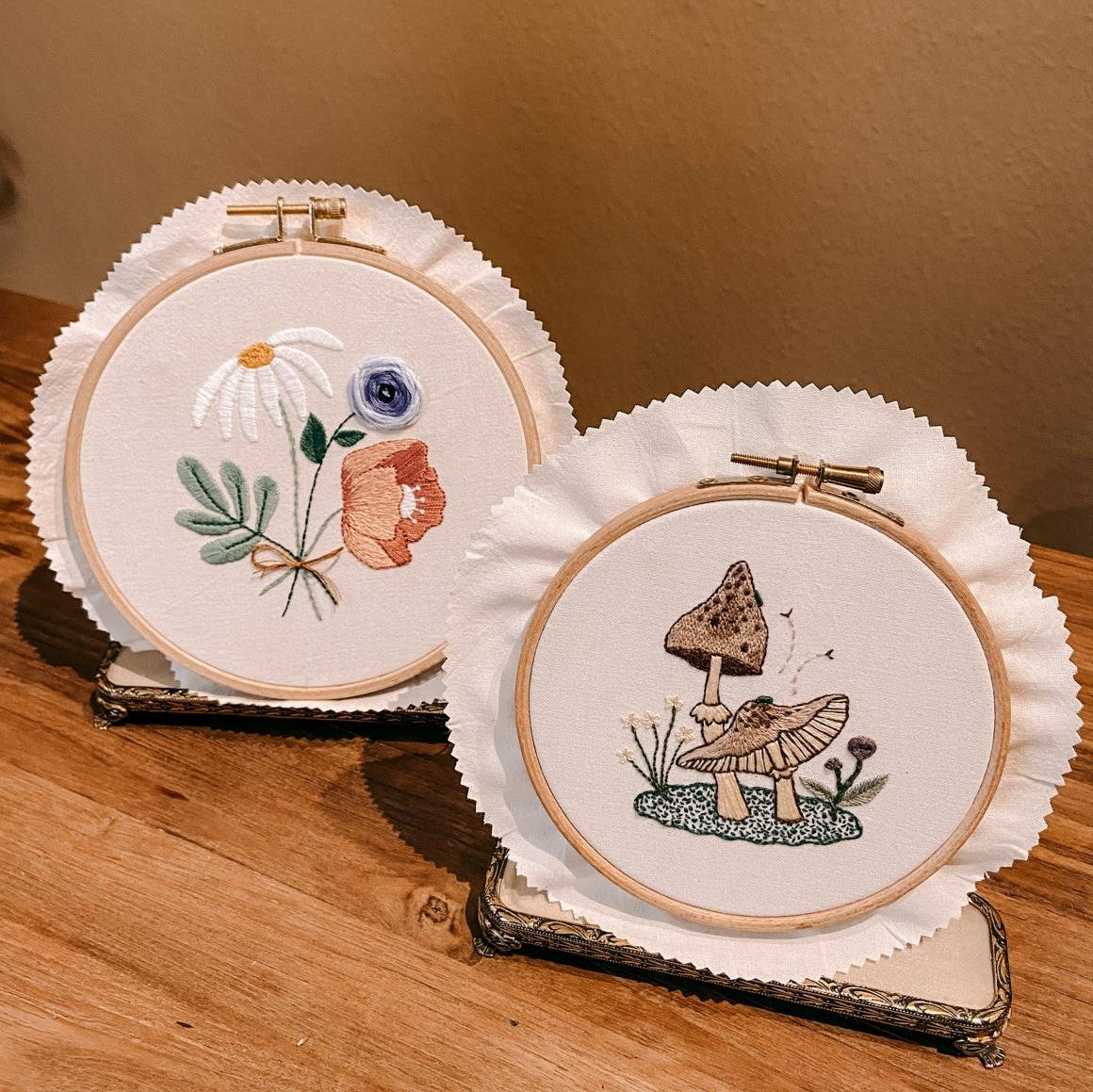 Zwei fertige Stickereien – Pilz und Blumen – auf Holztisch arrangiert.