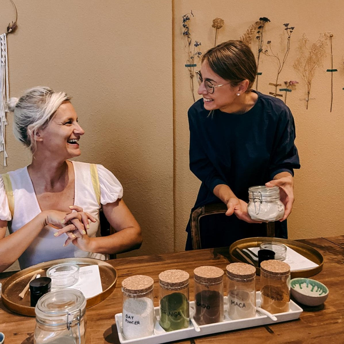 Zwei Frauen lachen vor Gläsern mit farbigen Pulvern – DIY Hautpflege Workshop.