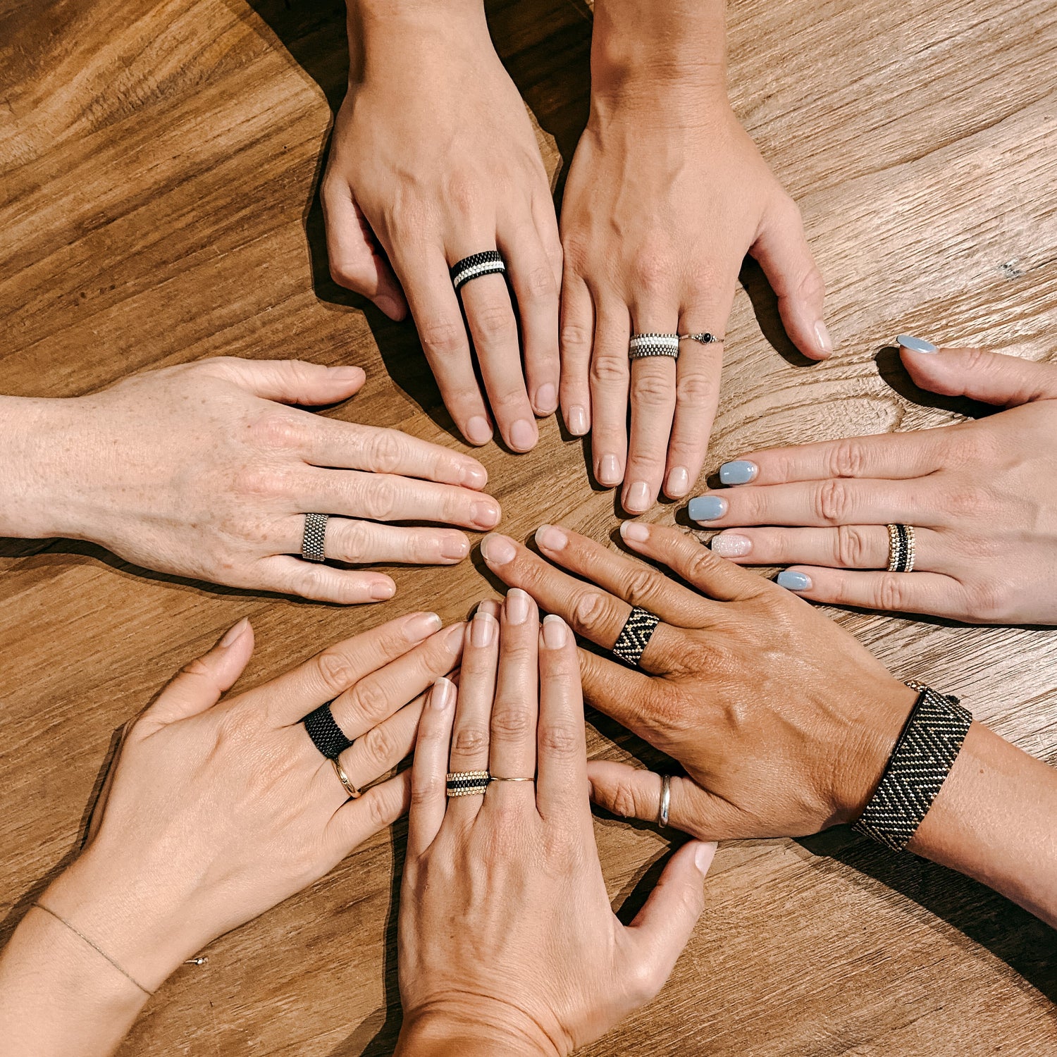 Sieben Hände mit verschiedenen Ringen auf Holztisch – Perlen Schmuck selber machen, Miyuki Perlen