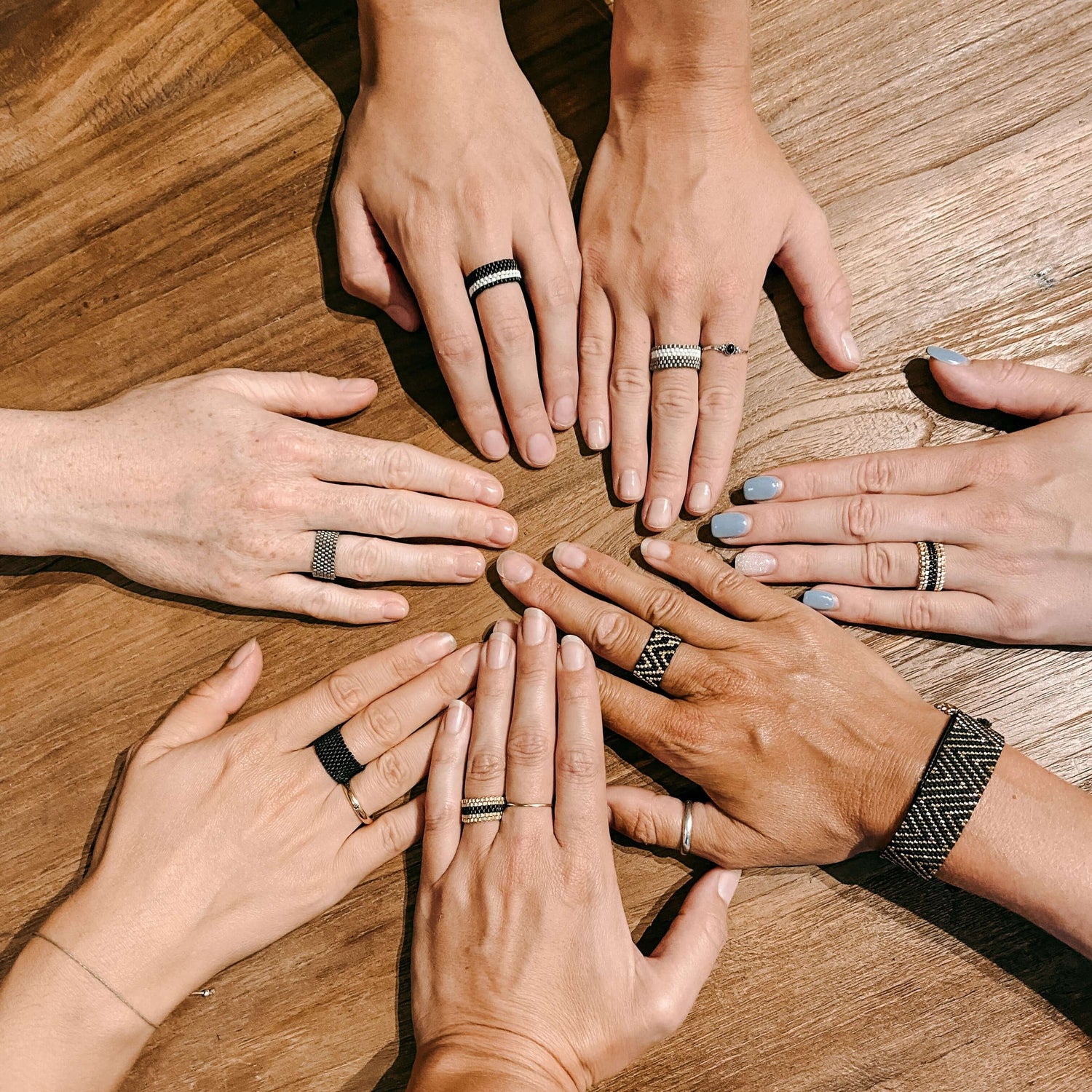 Sieben Hände mit verschiedenen Ringen auf Holztisch – Perlen Schmuck selber machen, Miyuki Perlen