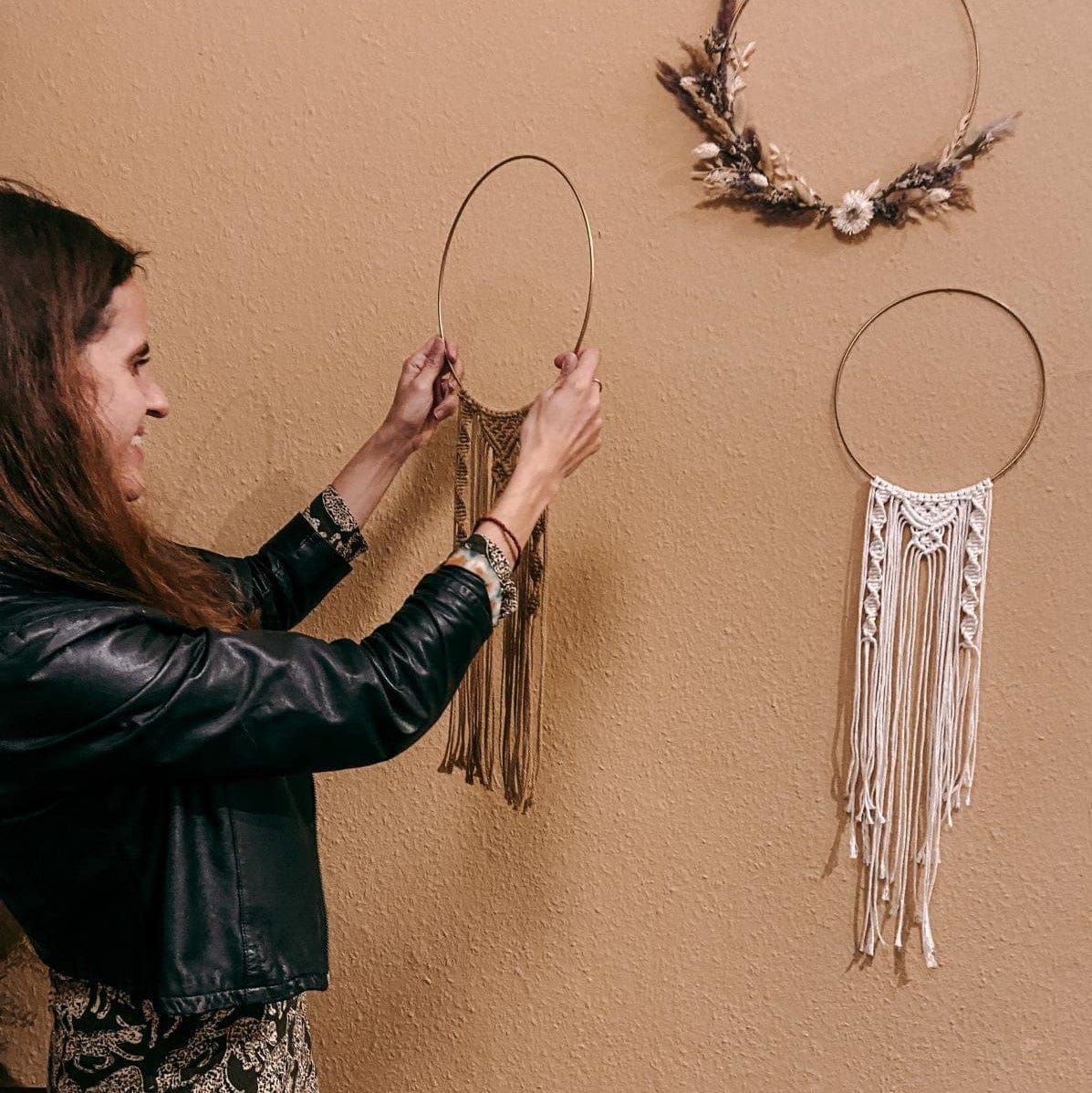 Frau hält fertigen Makramee-Ring in die Höhe, Wand mit Makramee im Hintergrund – Boho Flair, Makramee Kurs, Handarbeit.