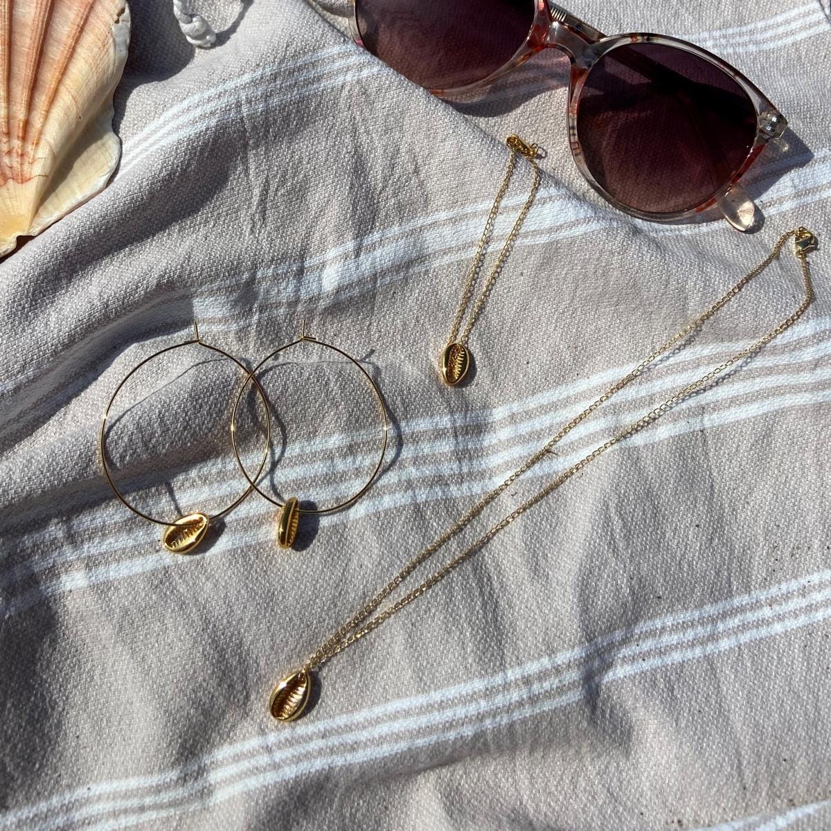 Kauri-Muschel Creolen und Kette auf Strandhandtuch mit Sonnenbrille arrangiert.