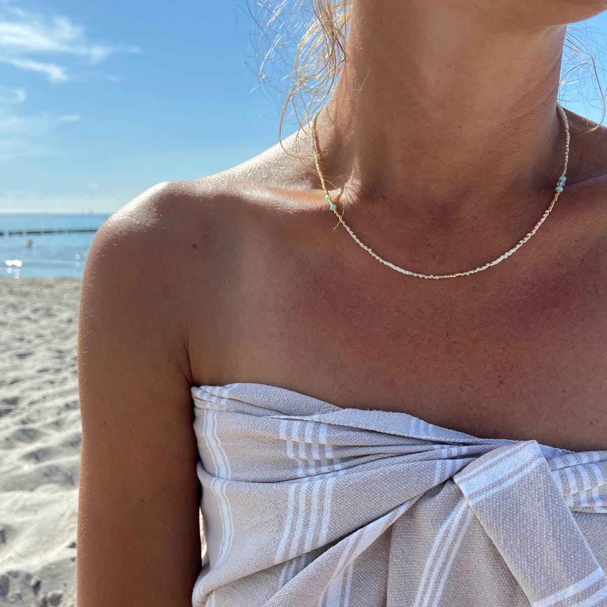 Person trägt Perlenkette „Summerfeeling“ am Hals am Strand in Mint, Weiß und Gold.