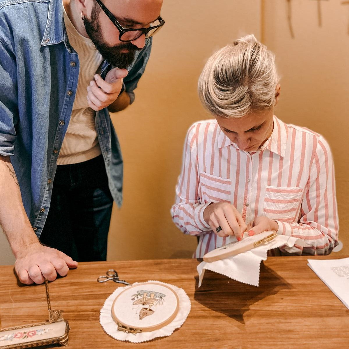 Zwei Personen bearbeiten Stickereien mit fertigem Pilzprojekt auf Tisch.