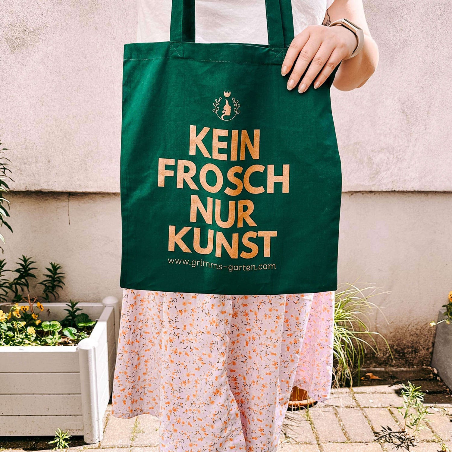 Person hält grünen Jutebeutel mit goldenem Print „KEIN FROSCH NUR KUNST“ von Grimms Garten in die Höhe.
