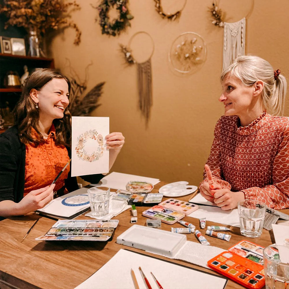 GRIMMS GARTEN Workshop – Frau hält Aquarell-Blumenkranz hoch, bunte Farben auf Tisch für kreatives Gestalten.