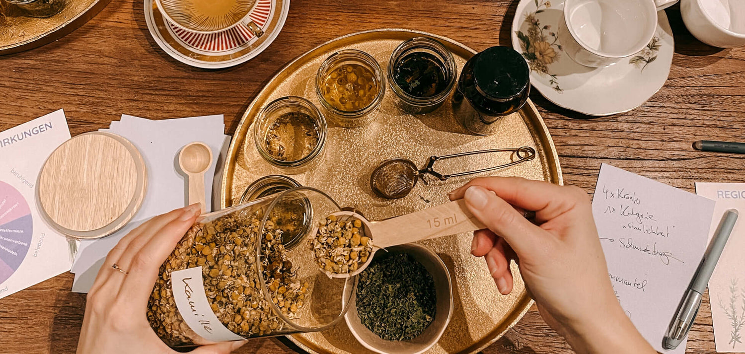 Goldenens Tablett mit Kräutern in Gläsern, Hände schöpfen Kamille – Kräutertee Workshop im GRIMMS GARTEN erleben.