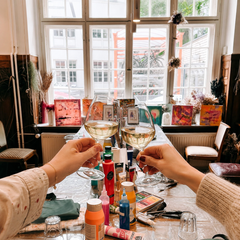 Vorbereiteter Tisch mit Acrylfarben, Pinseln und kreativen Materialien für den Abstraktes Malen Workshop von GRIMMS GARTEN Berlin. Im Vordergrund stoßen zwei Personen mit Wein an